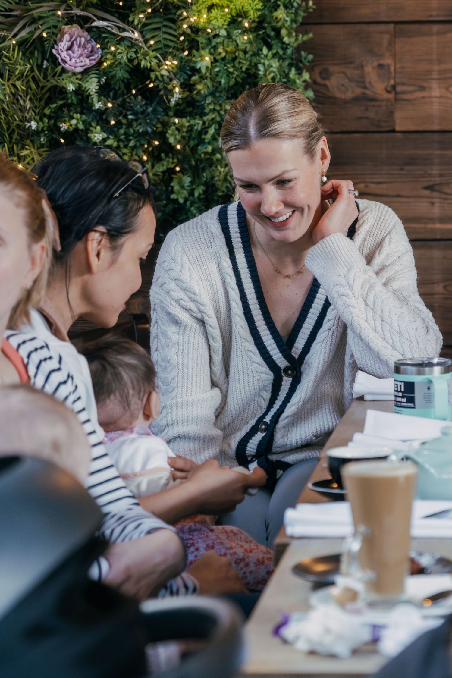 Group enjoying coffee and conversation at an outdoor cafe table with rustic wood wall and greenery in background.