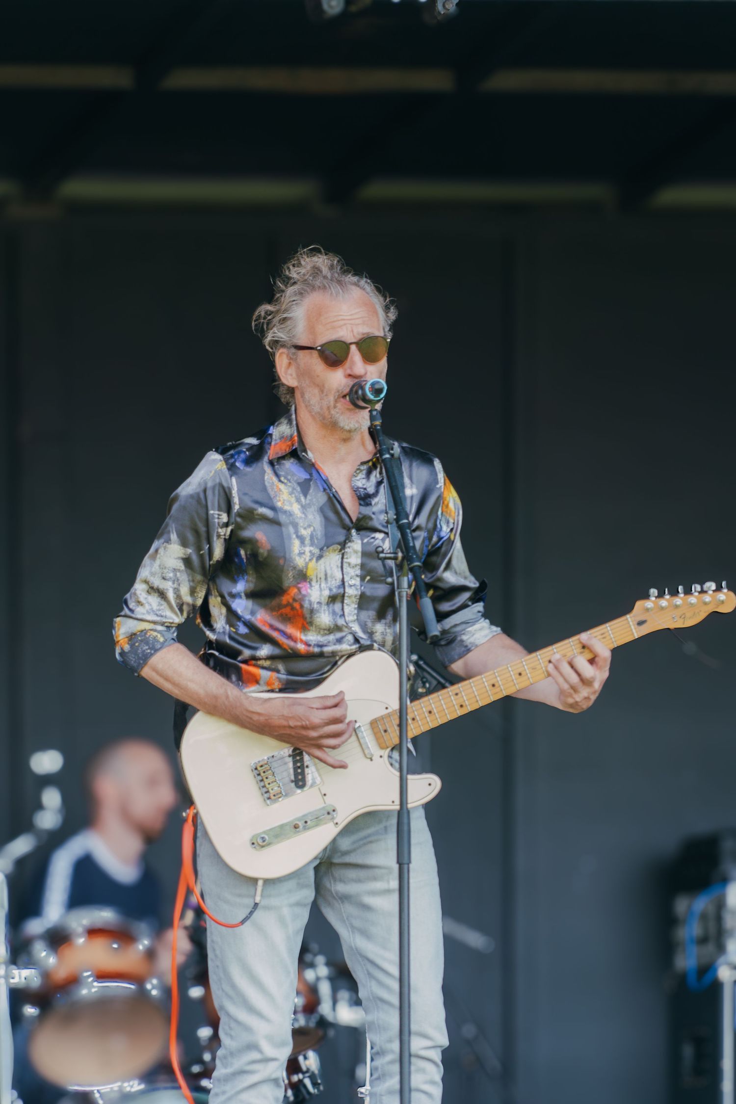 Musician in tie-dye shirt plays white Telecaster guitar on outdoor stage with drummer visible in background.