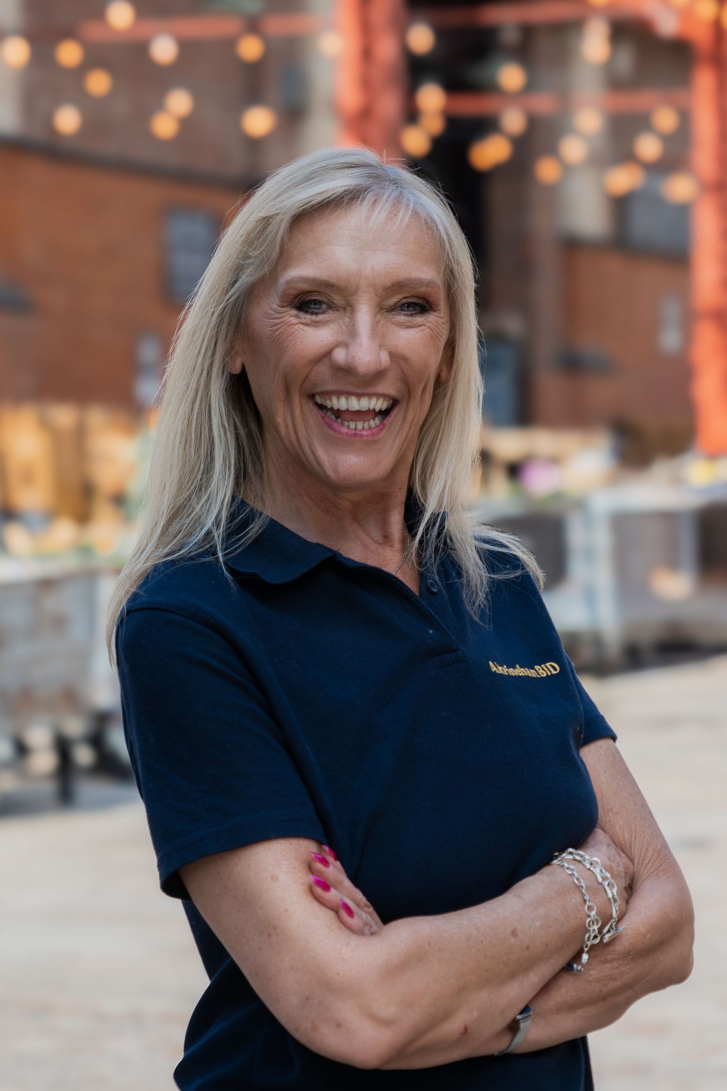 Professional wearing a navy blue polo shirt stands with crossed arms and a warm expression in a restaurant setting.