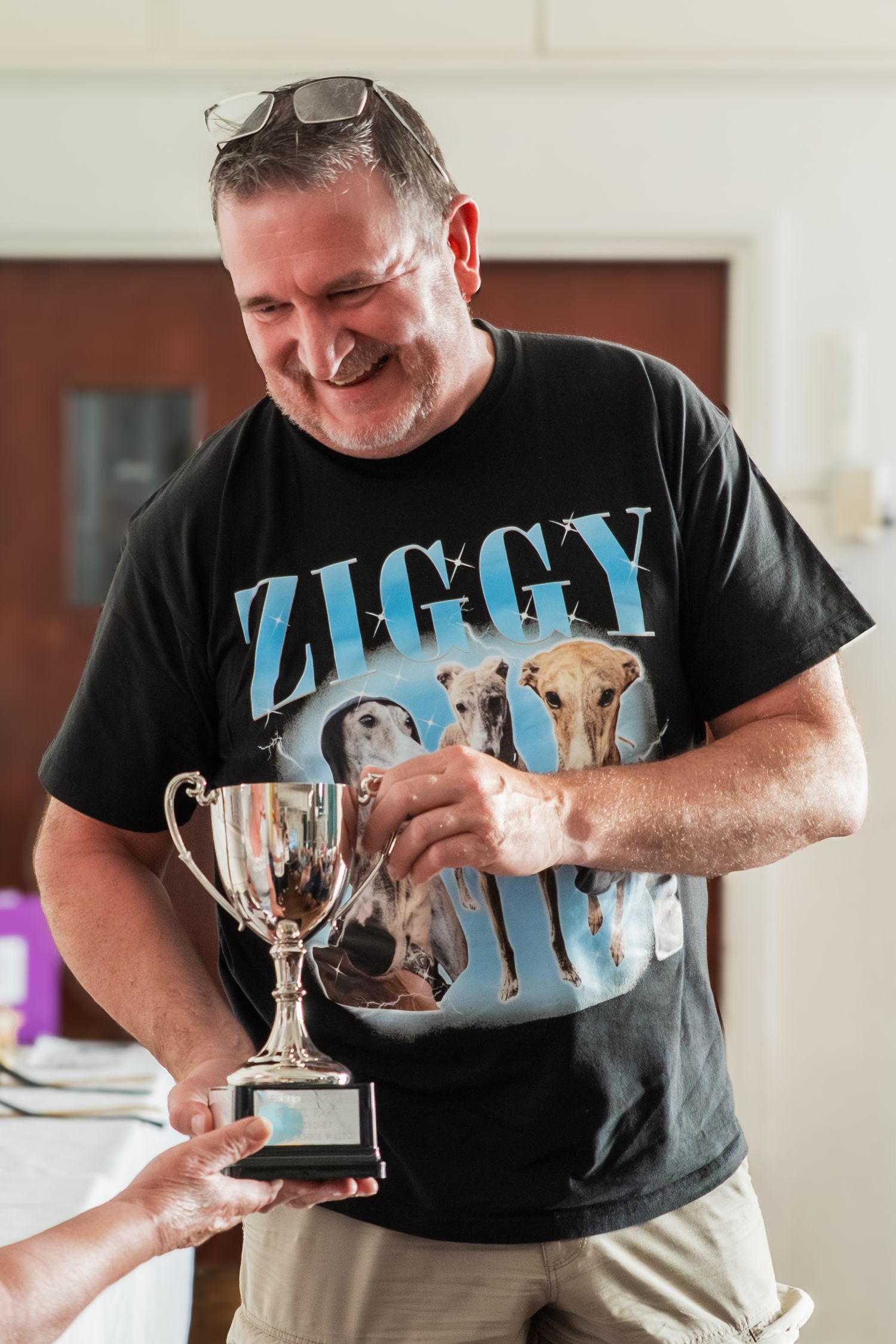 Someone wearing a Ziggy t-shirt smiles while receiving a trophy award indoors.