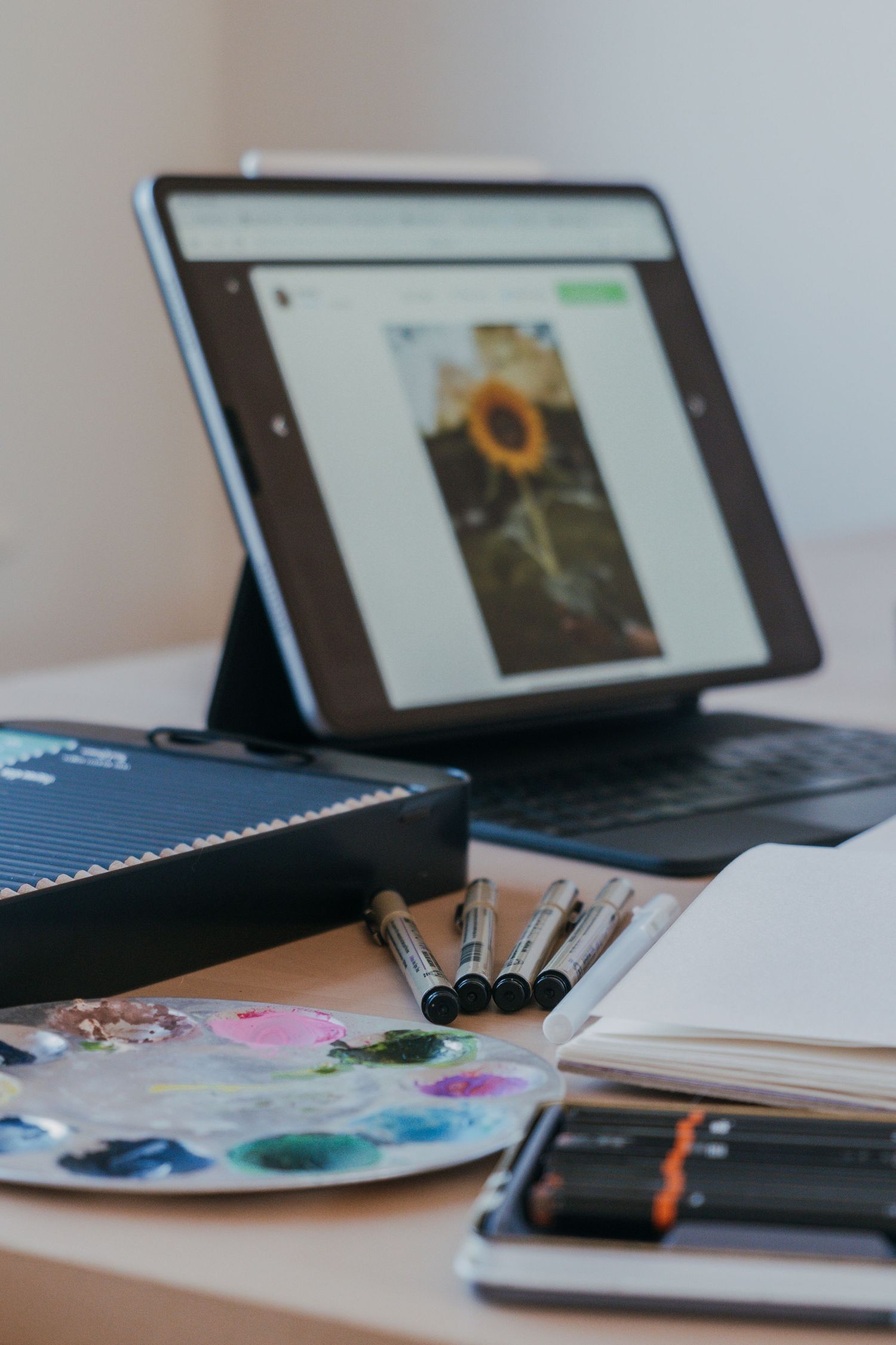Digital art workspace with tablet displaying sunflower artwork, paint palette, and drawing supplies on desk.