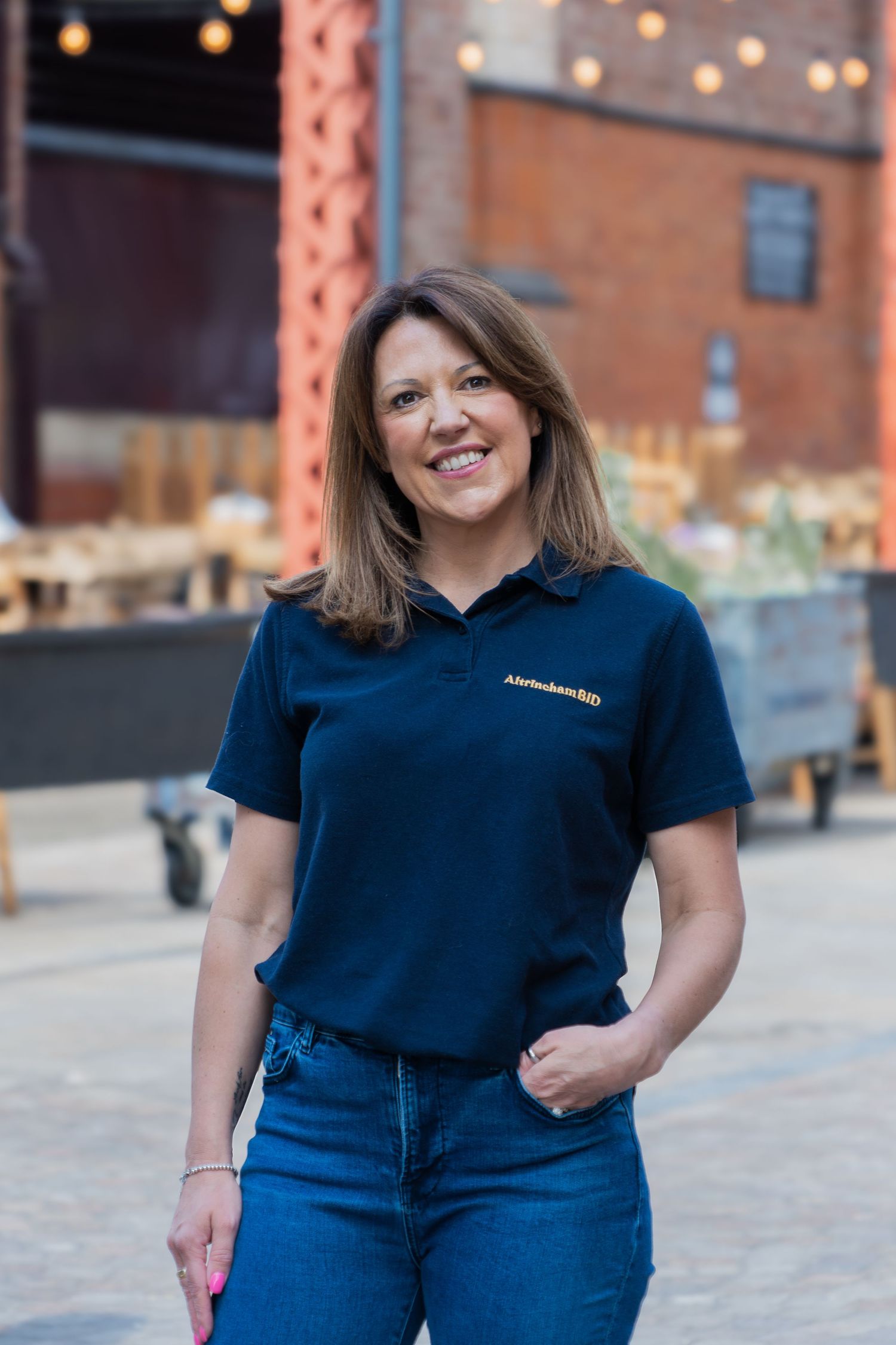 A person wearing a navy blue polo shirt and jeans poses outdoors against an industrial brick building with string lights.