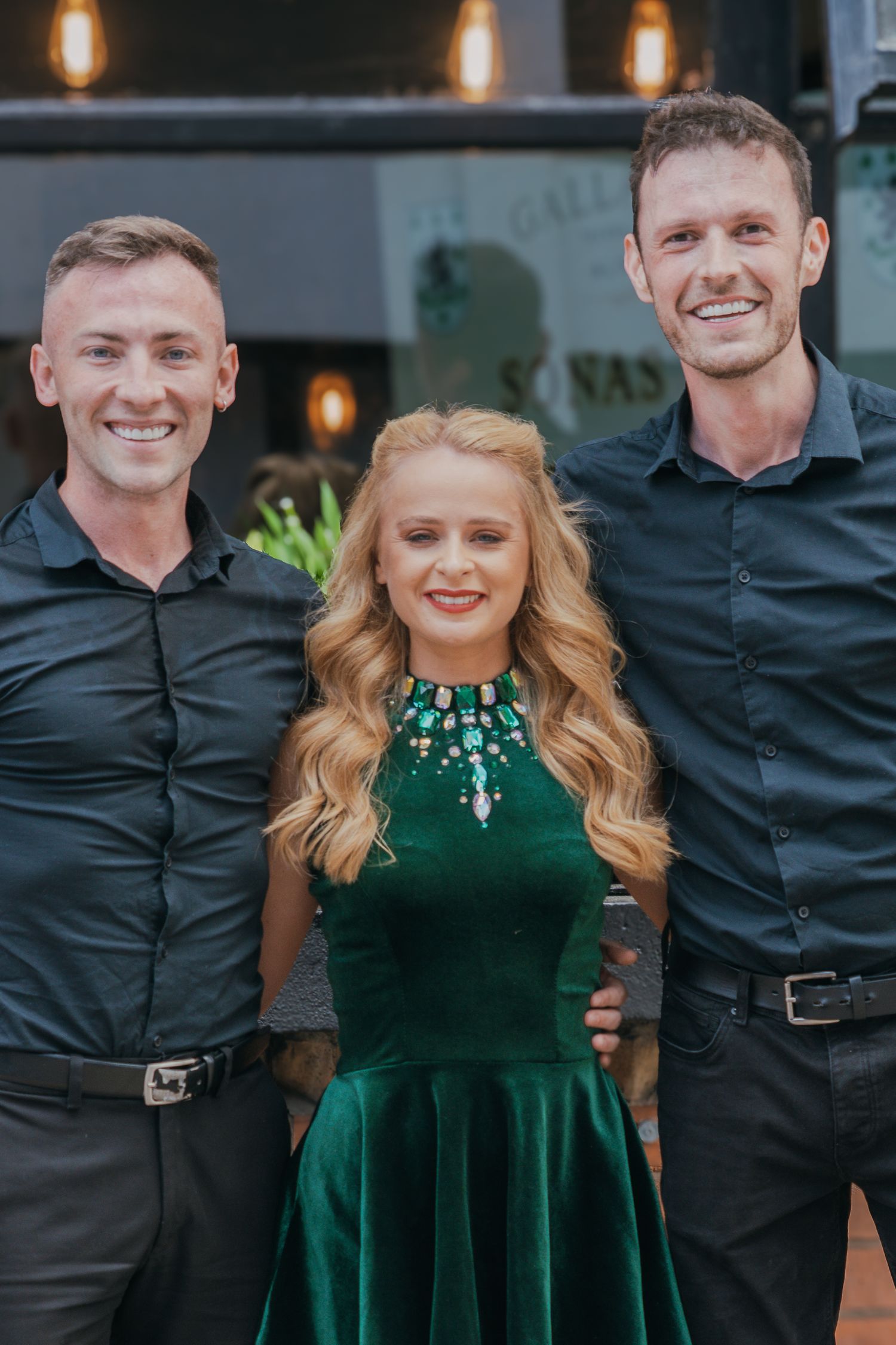 Three people dressed in formal attire pose together, with a woman in emerald velvet dress flanked by two men in dark shirts.
