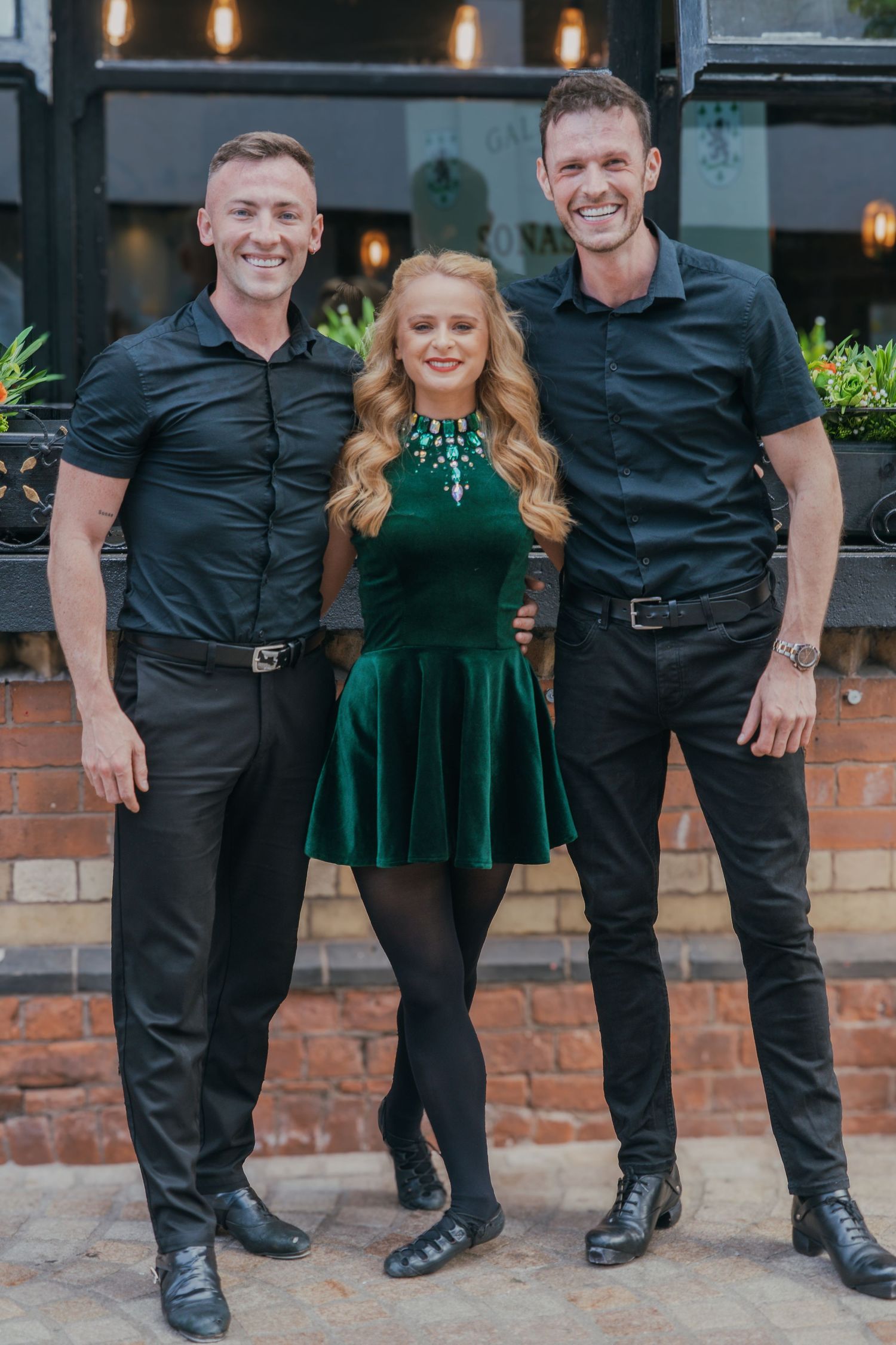 Three people pose together, with two wearing black shirts and one in an emerald green velvet dress.