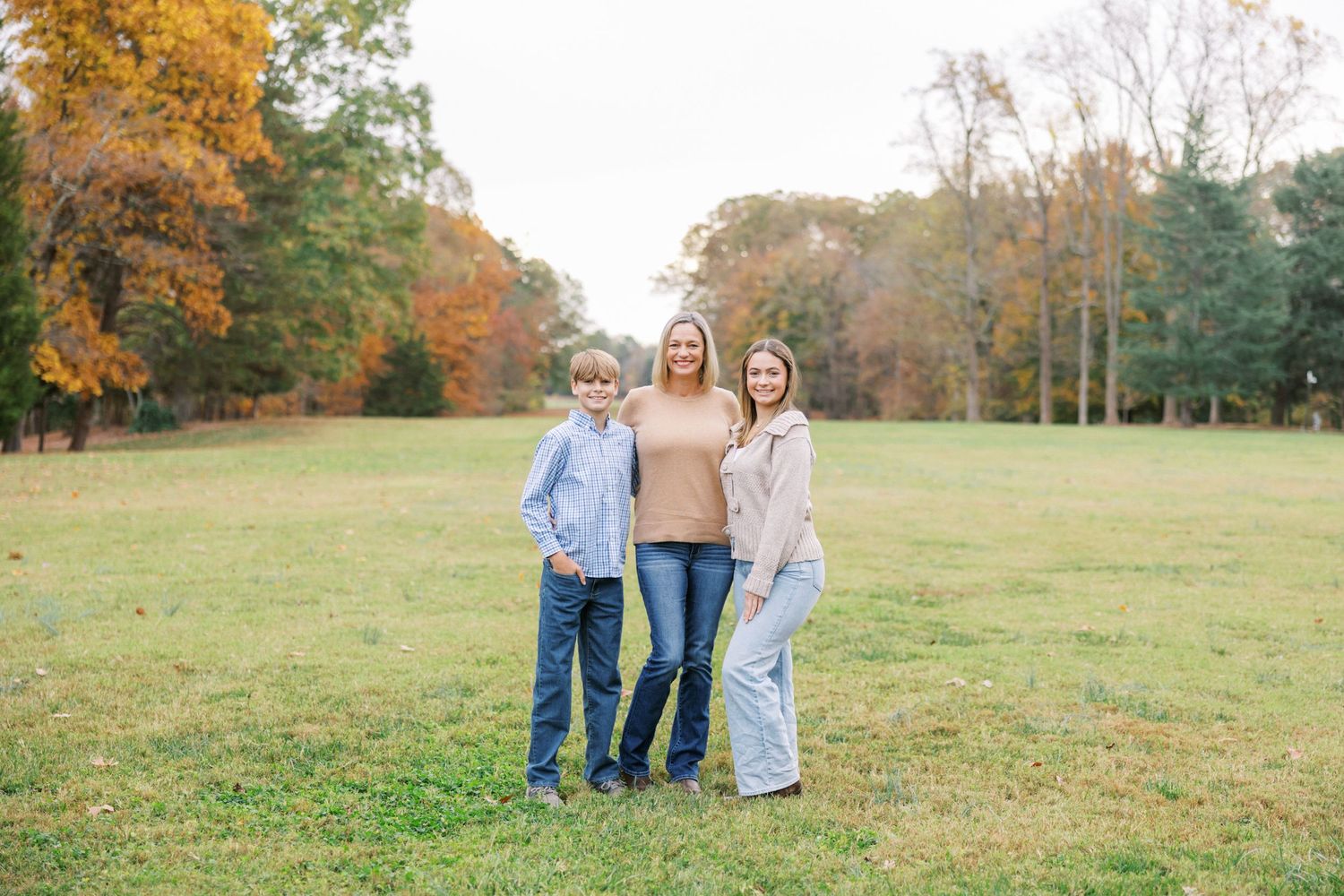 A group poses together in a grassy field surrounded by autumn trees with golden foliage on a sunny day.