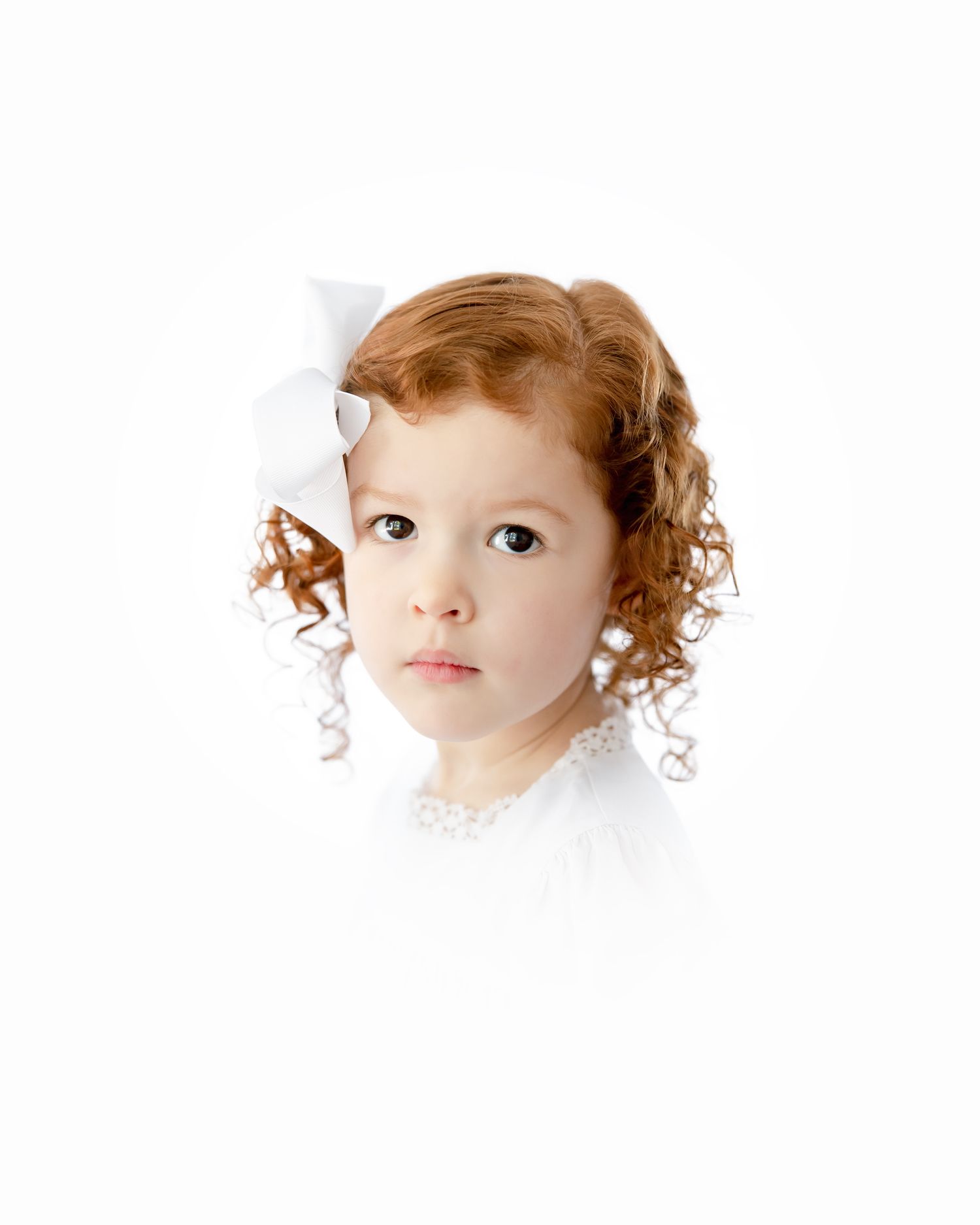 A child with red curly hair wearing white against a white background, looking at the camera with an earnest expression.