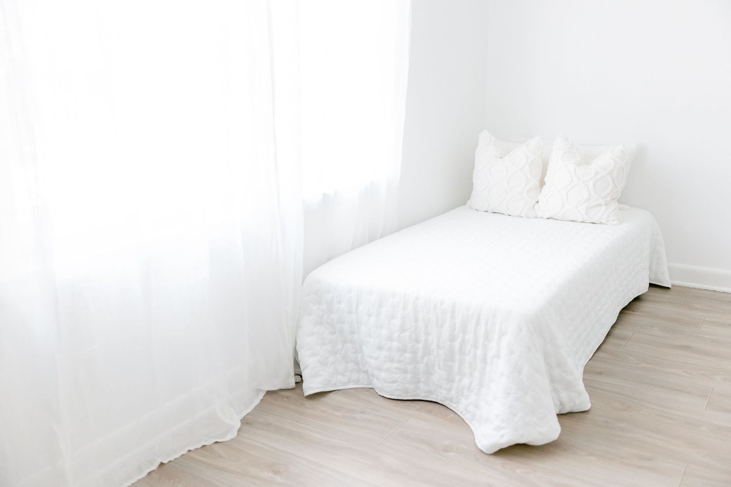 A minimalist white bed with quilted bedding sits against a white wall in a bright, airy bedroom with wooden flooring.
