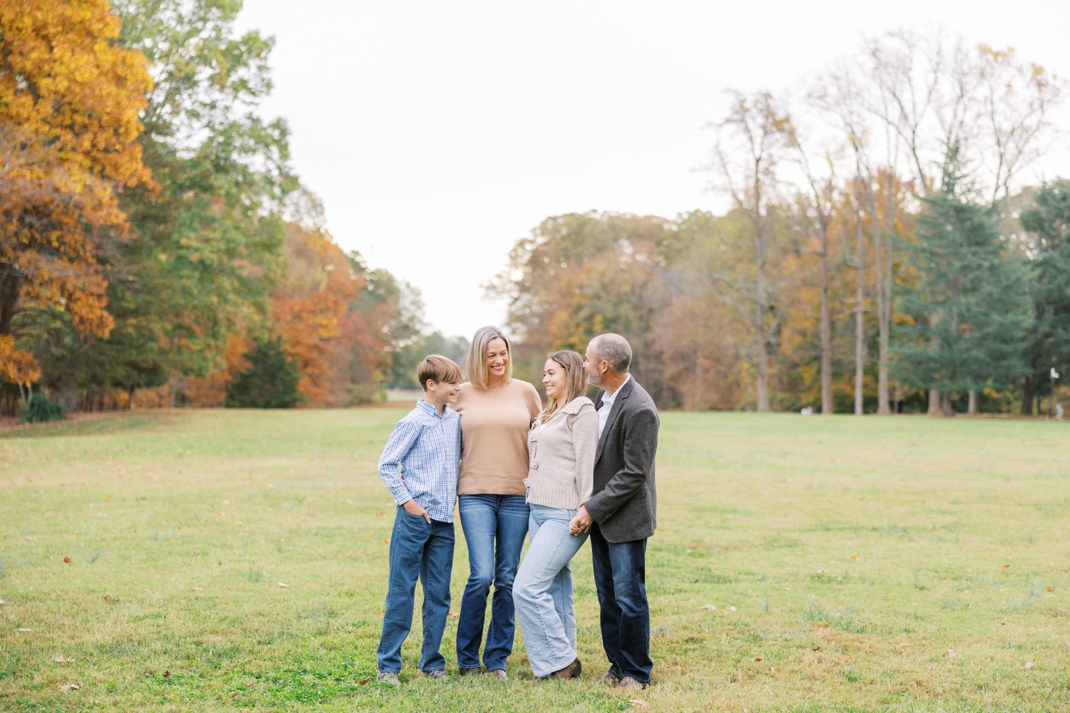 Group of people standing together outdoors on green grass during autumn with colorful fall foliage in background.