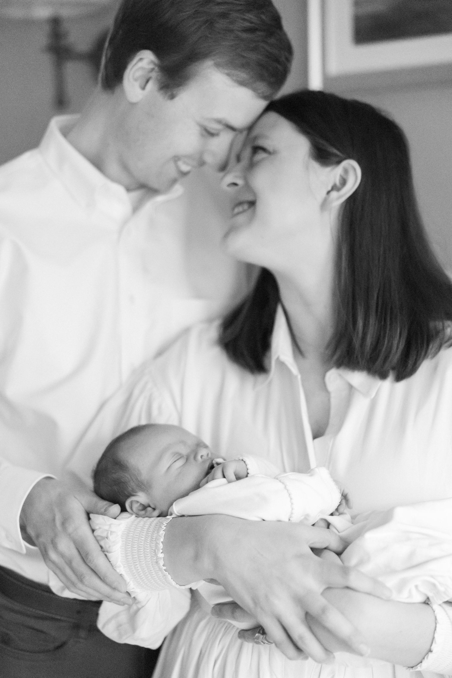 An intimate black and white photo of a couple sharing a tender moment while holding their sleeping newborn.