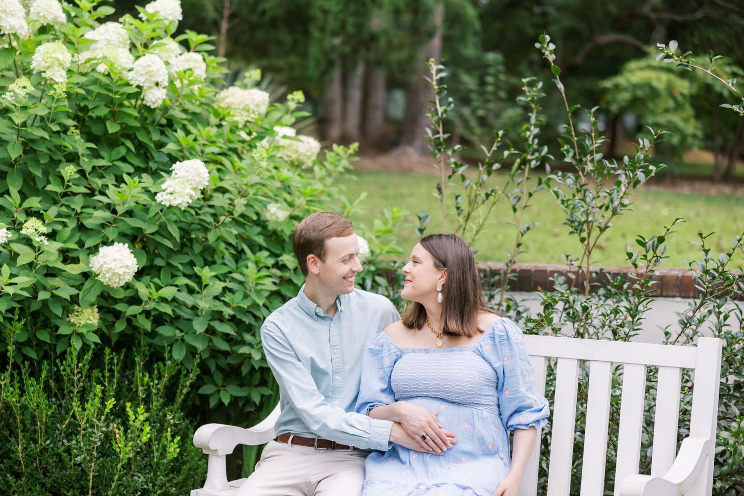A couple sits together on a white garden bench surrounded by blooming hydrangeas and greenery.