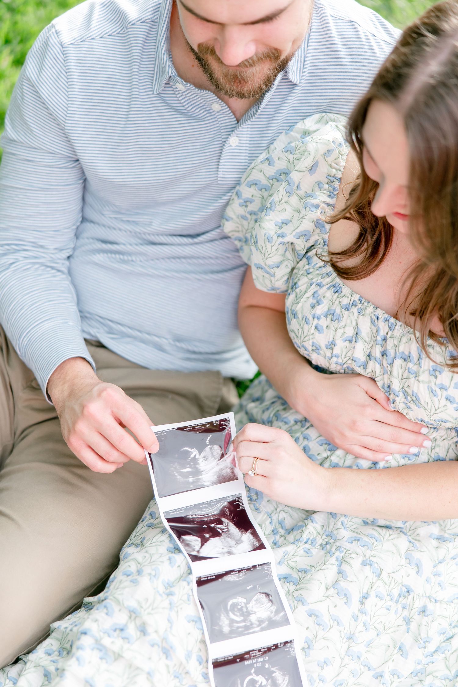 A couple in casual attire sits together outdoors looking at ultrasound photos they're holding.