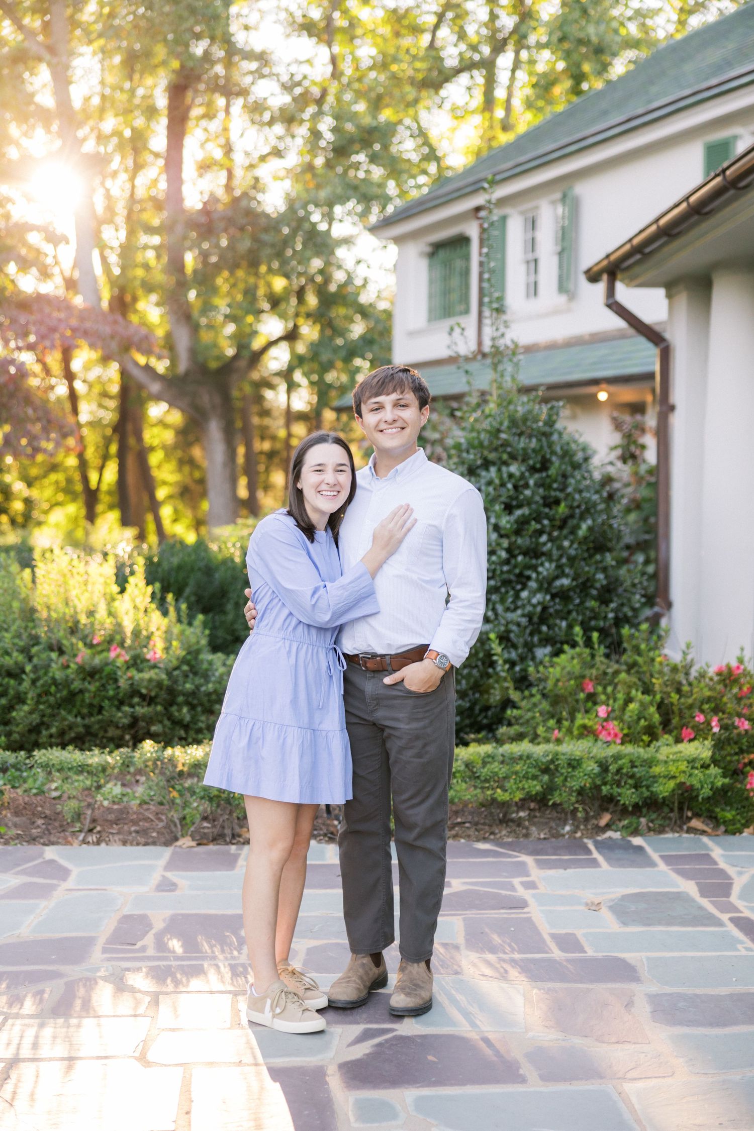 A couple poses together on a stone patio during golden hour with sunlight filtering through trees.