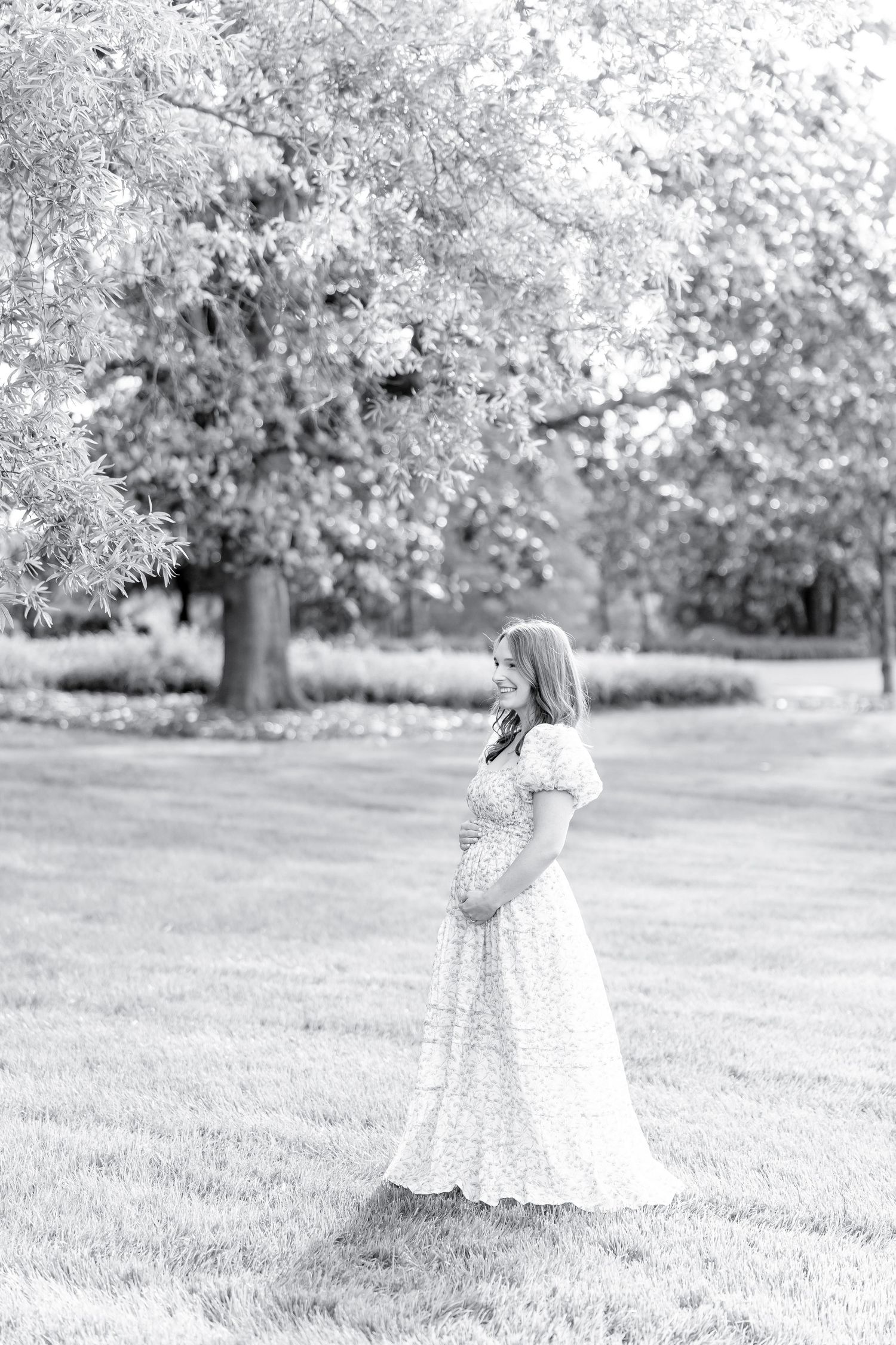A person in a white floral maxi dress stands in a park setting with blooming trees on a sunny day.