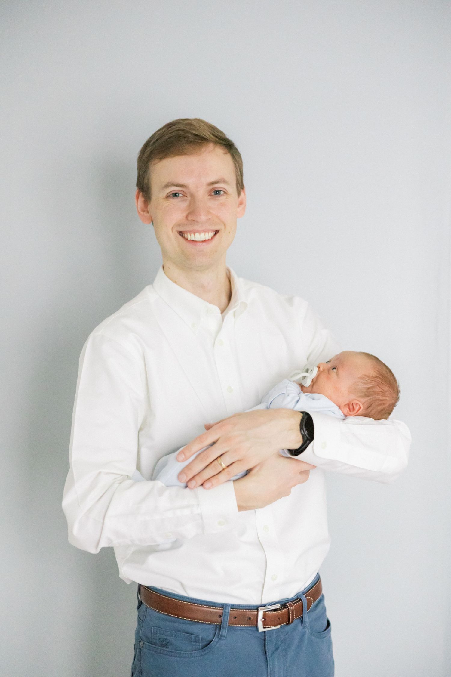 A person in a white shirt and jeans cradles a sleeping newborn baby against a gray wall.