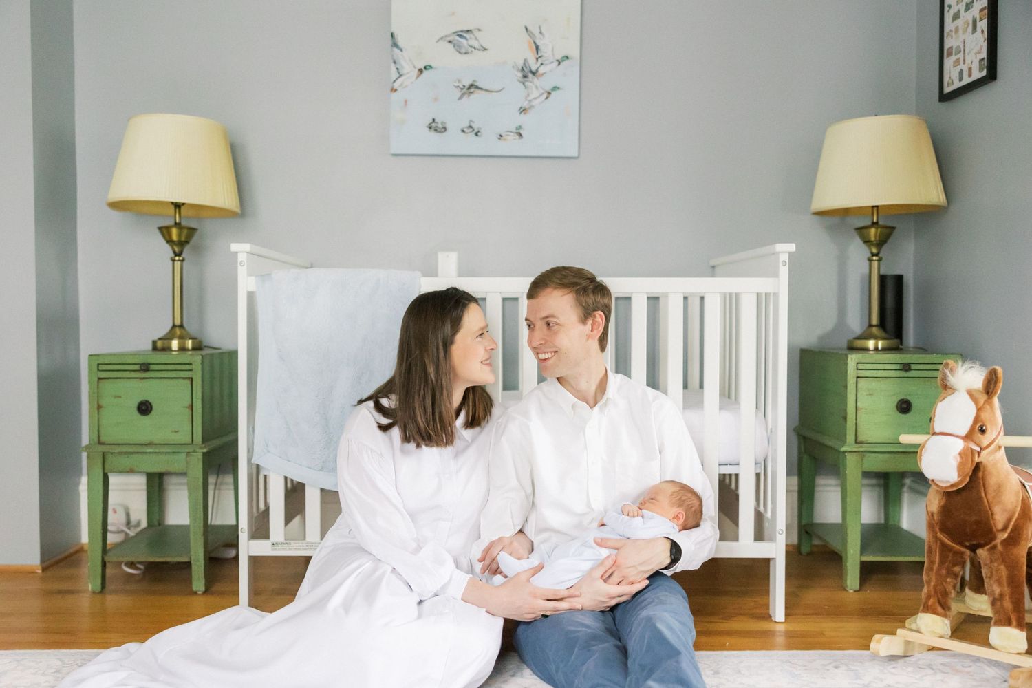 A couple sits next to a white crib with matching green nightstands and lamps in a cozy neutral-toned nursery.