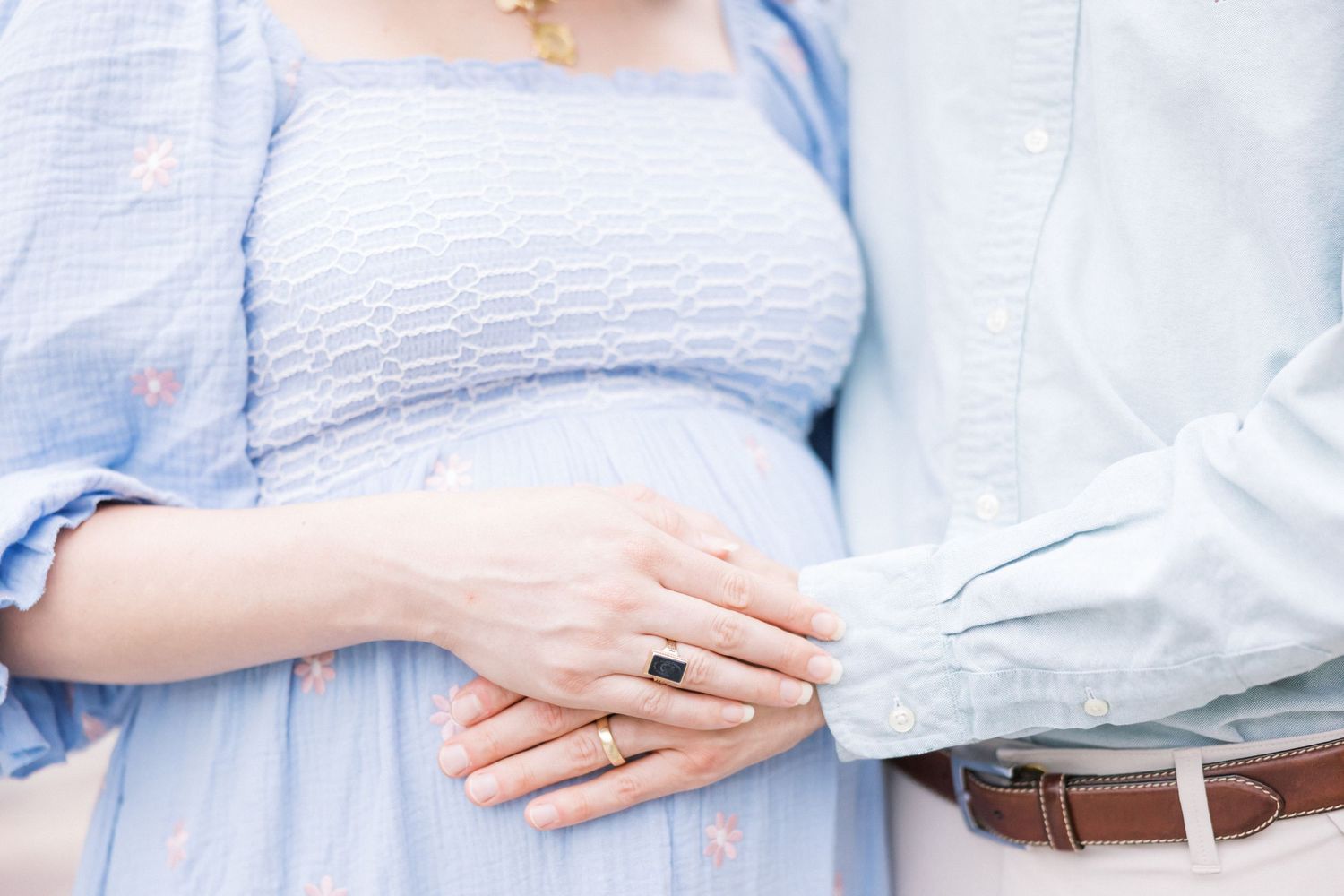 Close-up detail of hands resting on a baby bump while wearing light blue clothing.