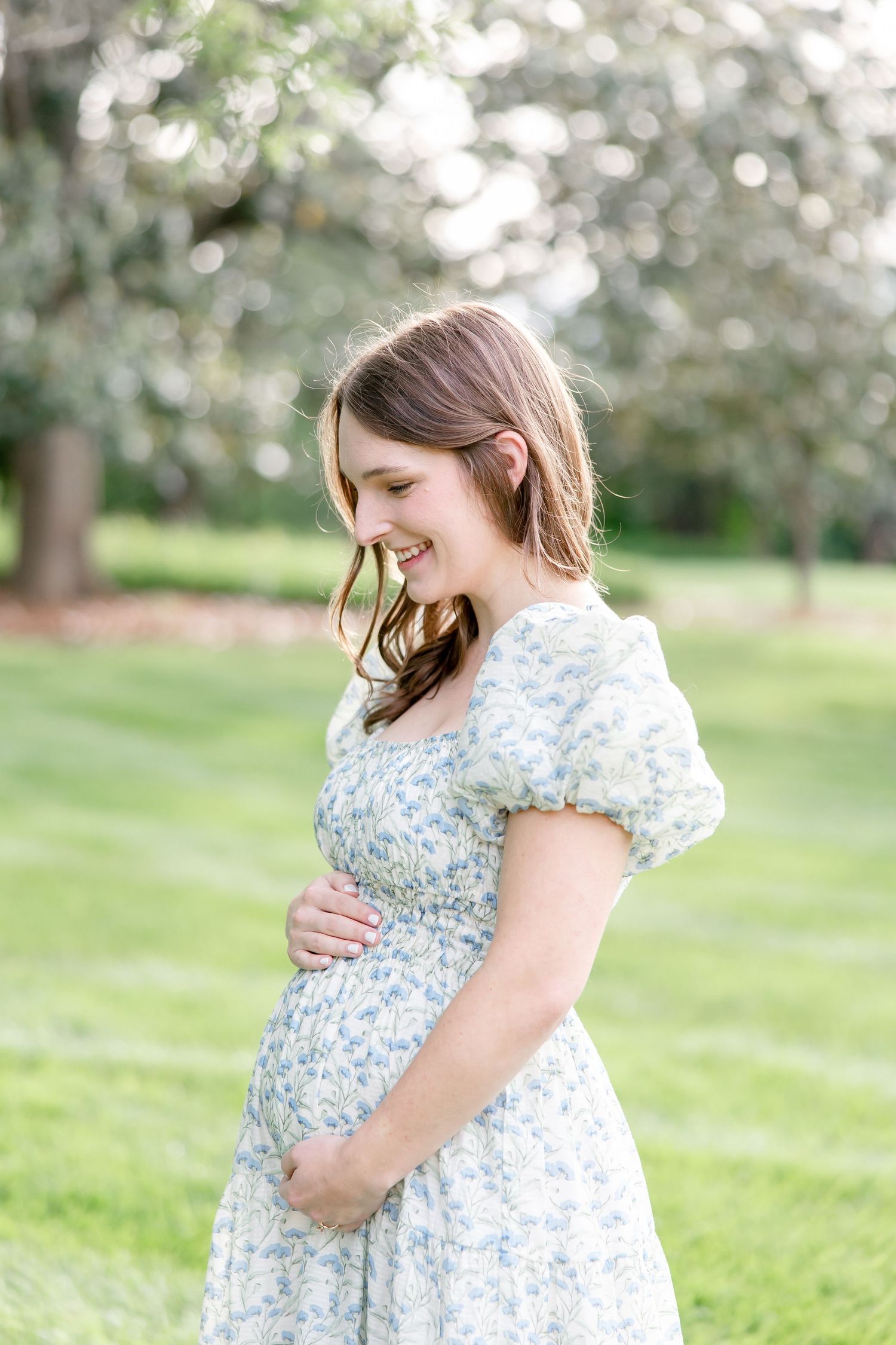 Expecting mother in floral dress smiles while standing in sunlit garden with soft bokeh background effect.