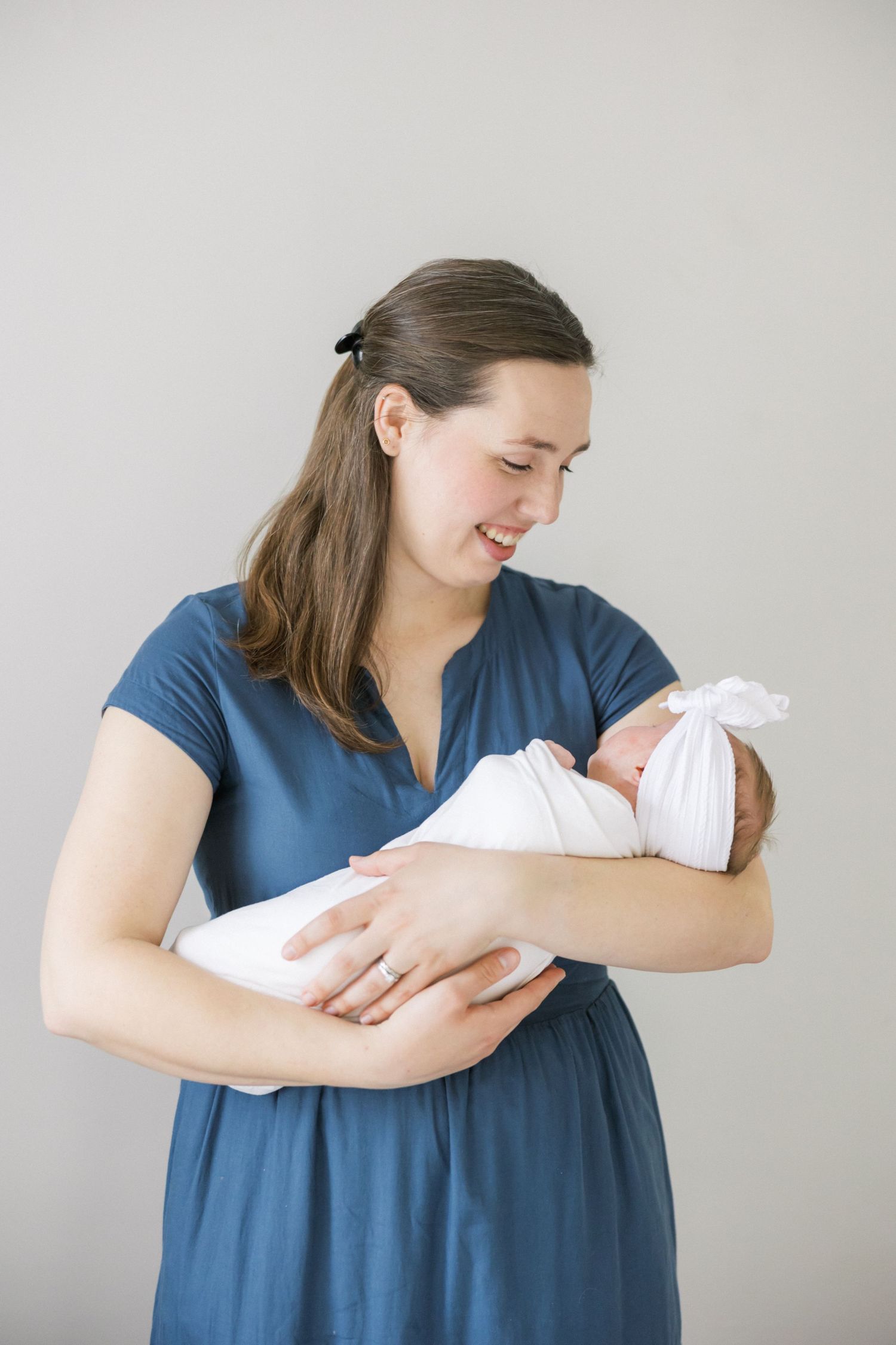 Woman in blue dress smiles lovingly while holding swaddled infant against gray background.
