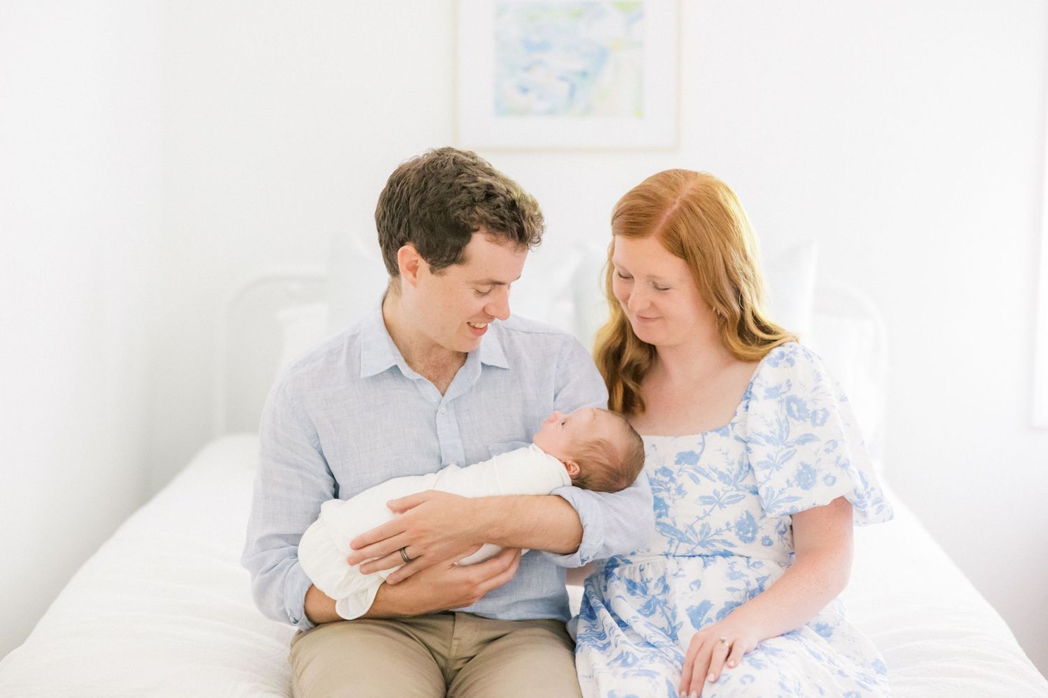 Two people sit on a white bed together, sharing a tender moment with a newborn baby wrapped in white.