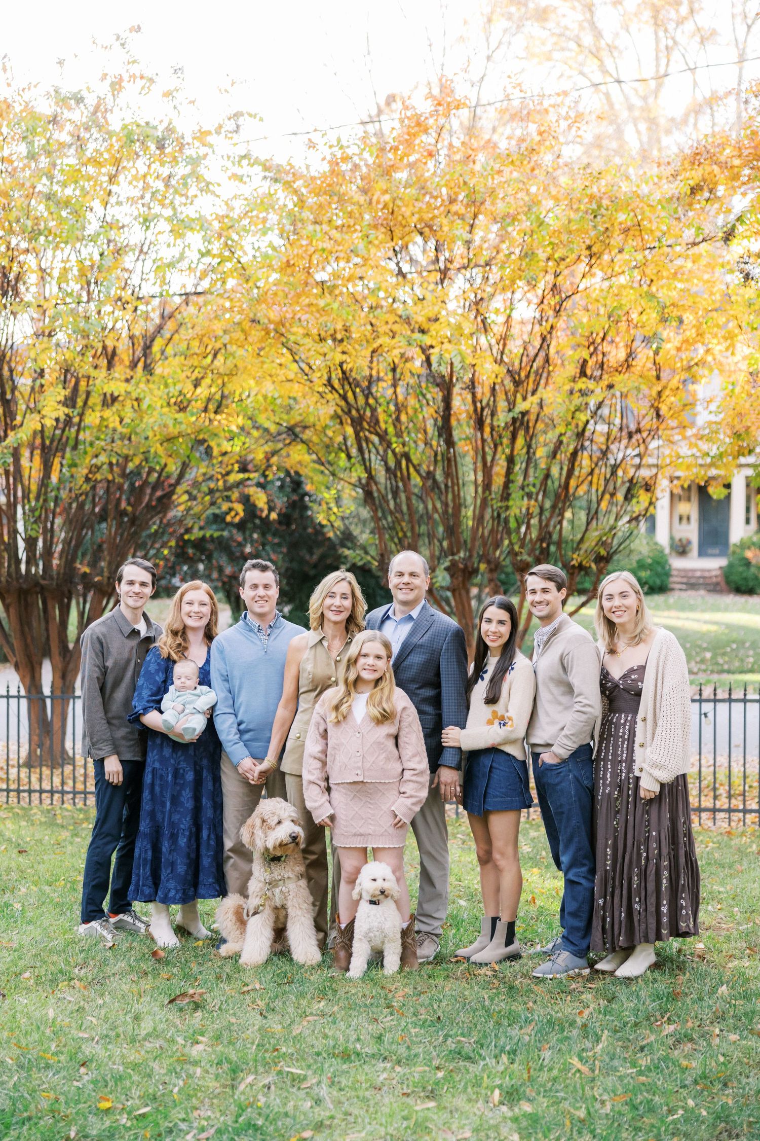 Large family group portrait outdoors with two goldendoodle dogs against autumn trees.