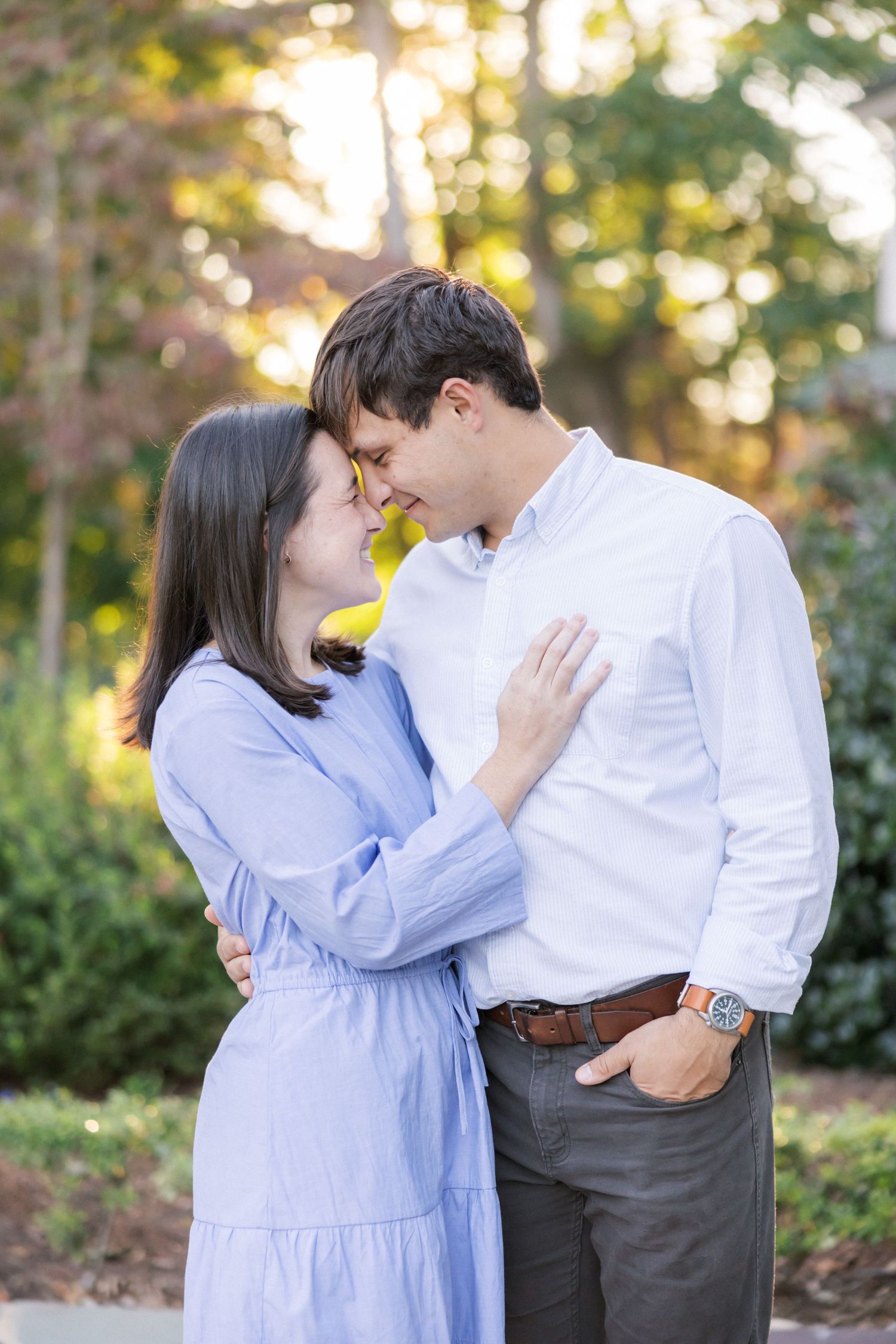 A couple embraces tenderly in a garden with golden sunlight filtering through the trees behind them.