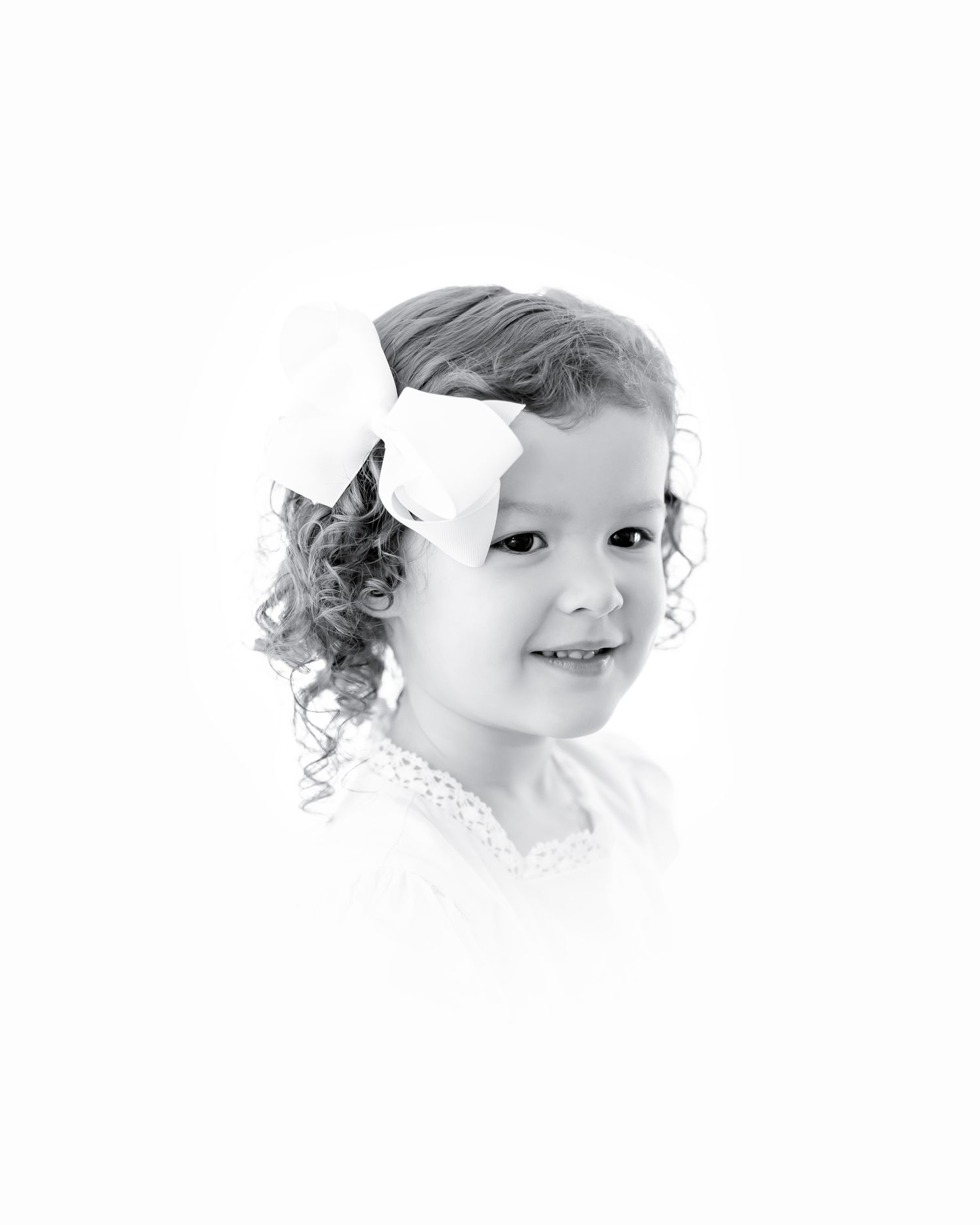 Black and white portrait of a smiling toddler with curls wearing a white bow and lace collar.
