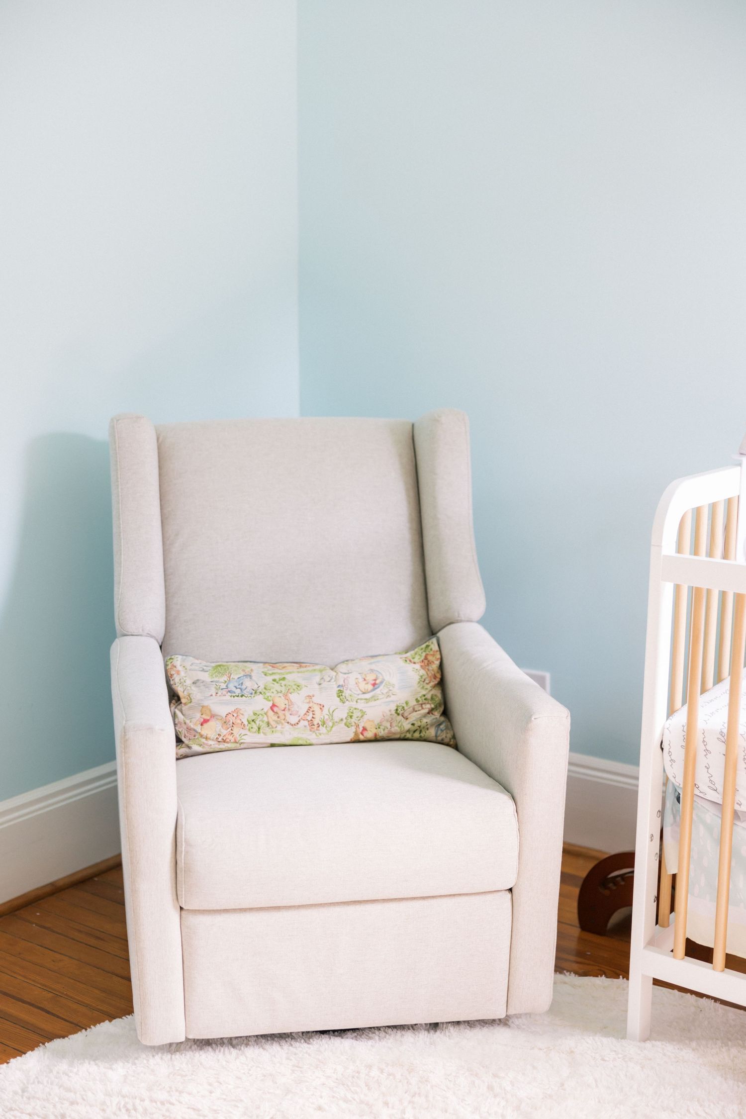 Cream-colored upholstered glider chair with floral pillow beside white crib in nursery.