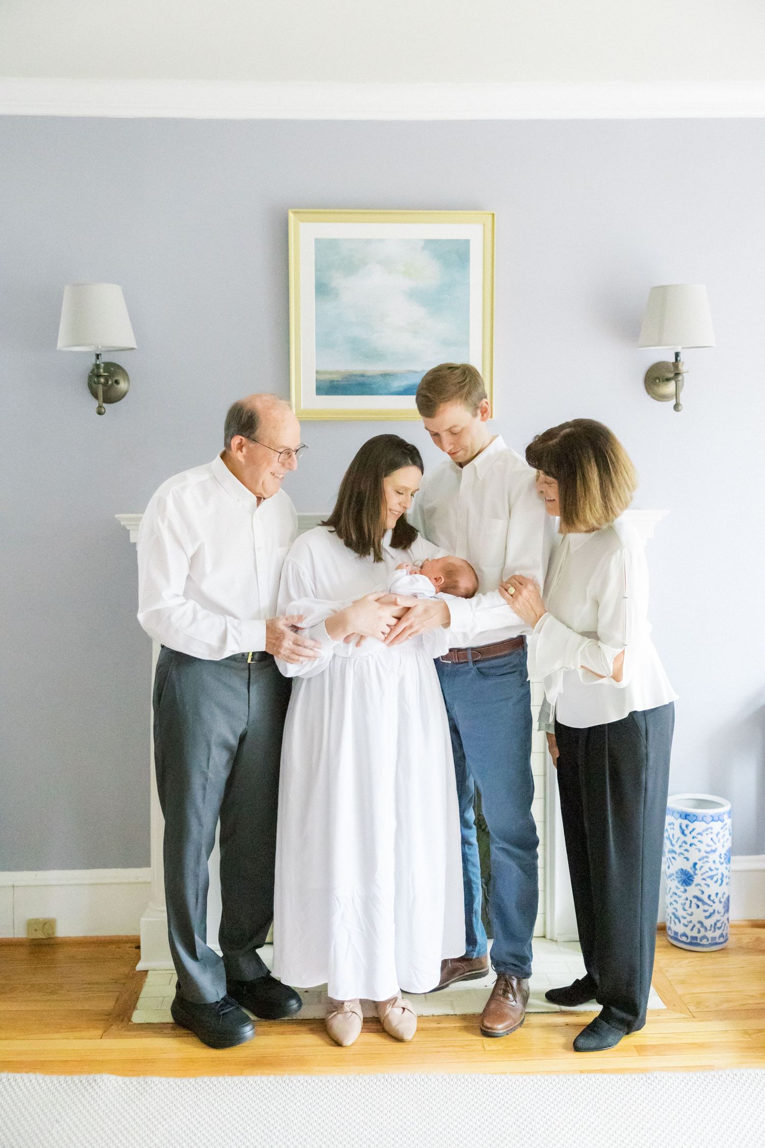 A family gathering in a living room to admire a newborn baby in front of coastal artwork and wall sconces.