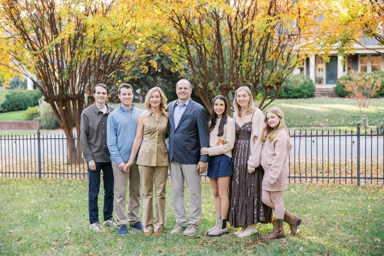 A multi-generational family poses together on a lawn with fall foliage and an iron fence in the background.