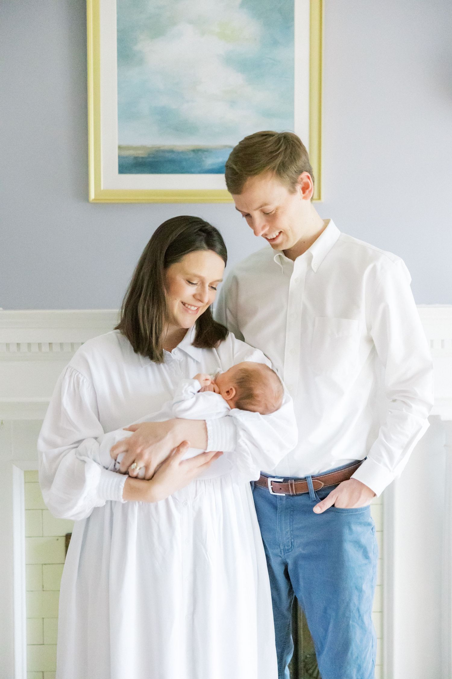 A couple stands together in a room with a seascape painting, gazing at their newborn baby.