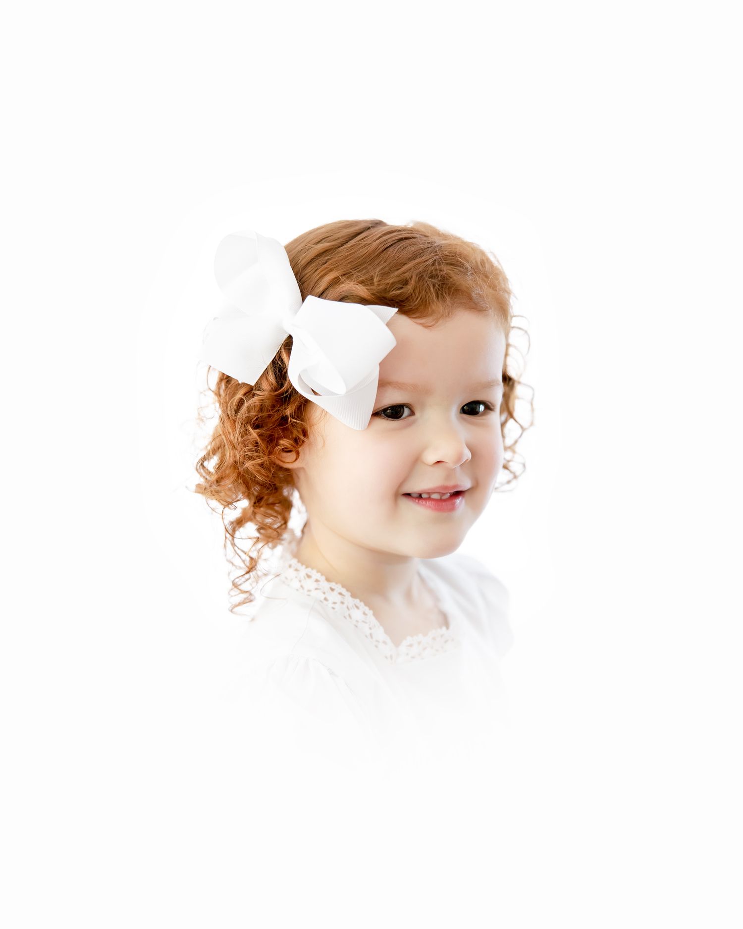An adorable child with red curly hair and a white bow wearing a lace collar smiles gently against a light background.