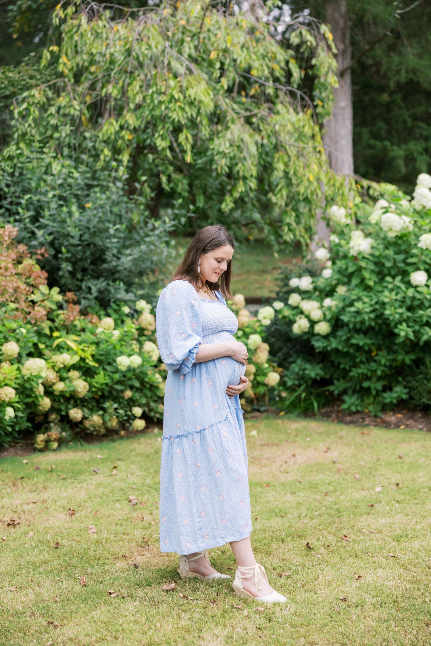 Woman in a light blue dress stands in a garden setting surrounded by white hydrangeas and trees.