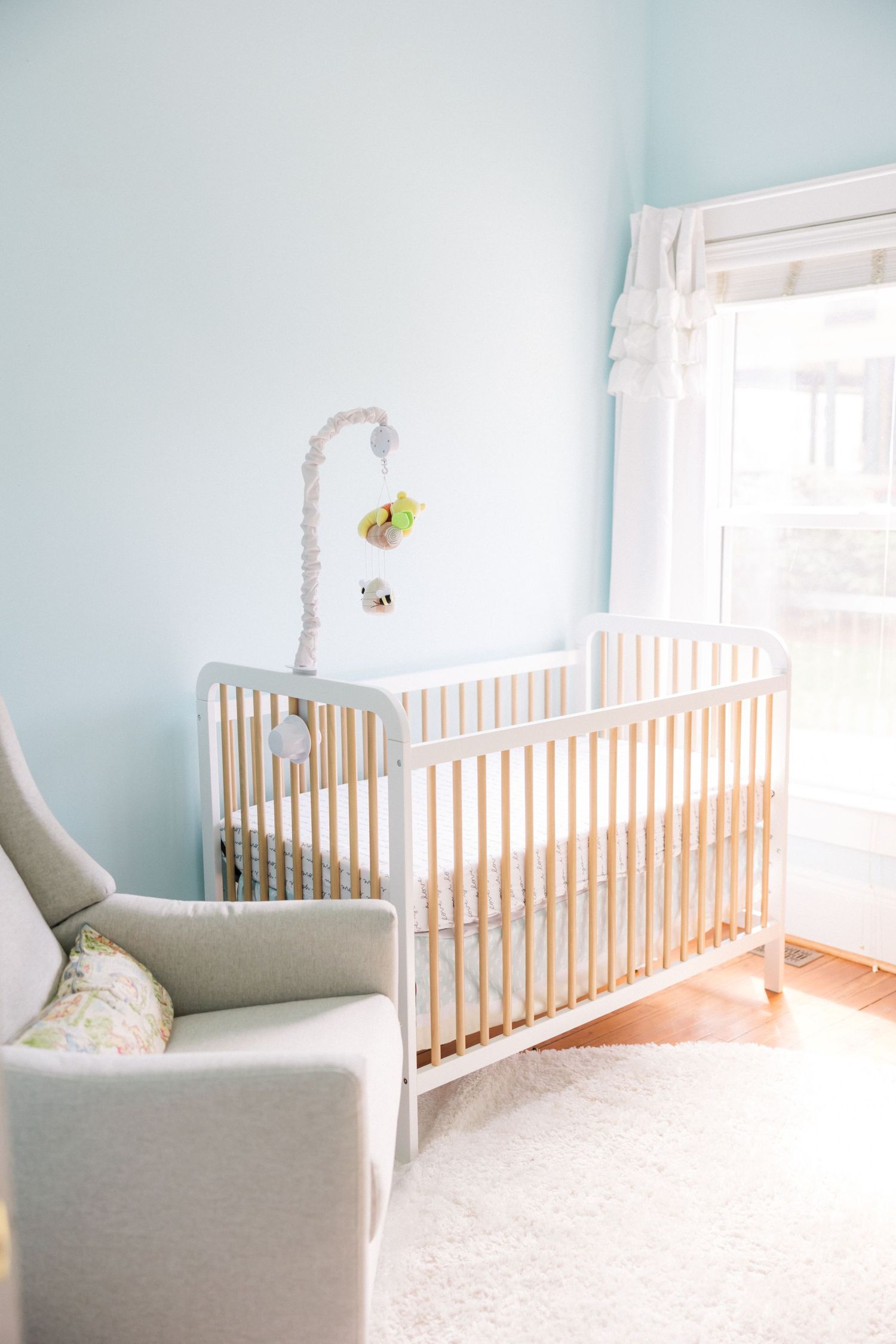 Modern white crib and gray armchair in minimalist nursery with sheer curtains.