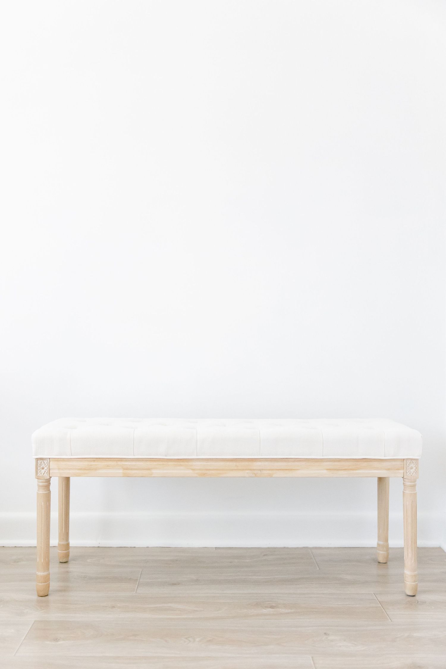 White upholstered wooden bench with elegant turned legs placed against a plain white wall on light hardwood flooring.
