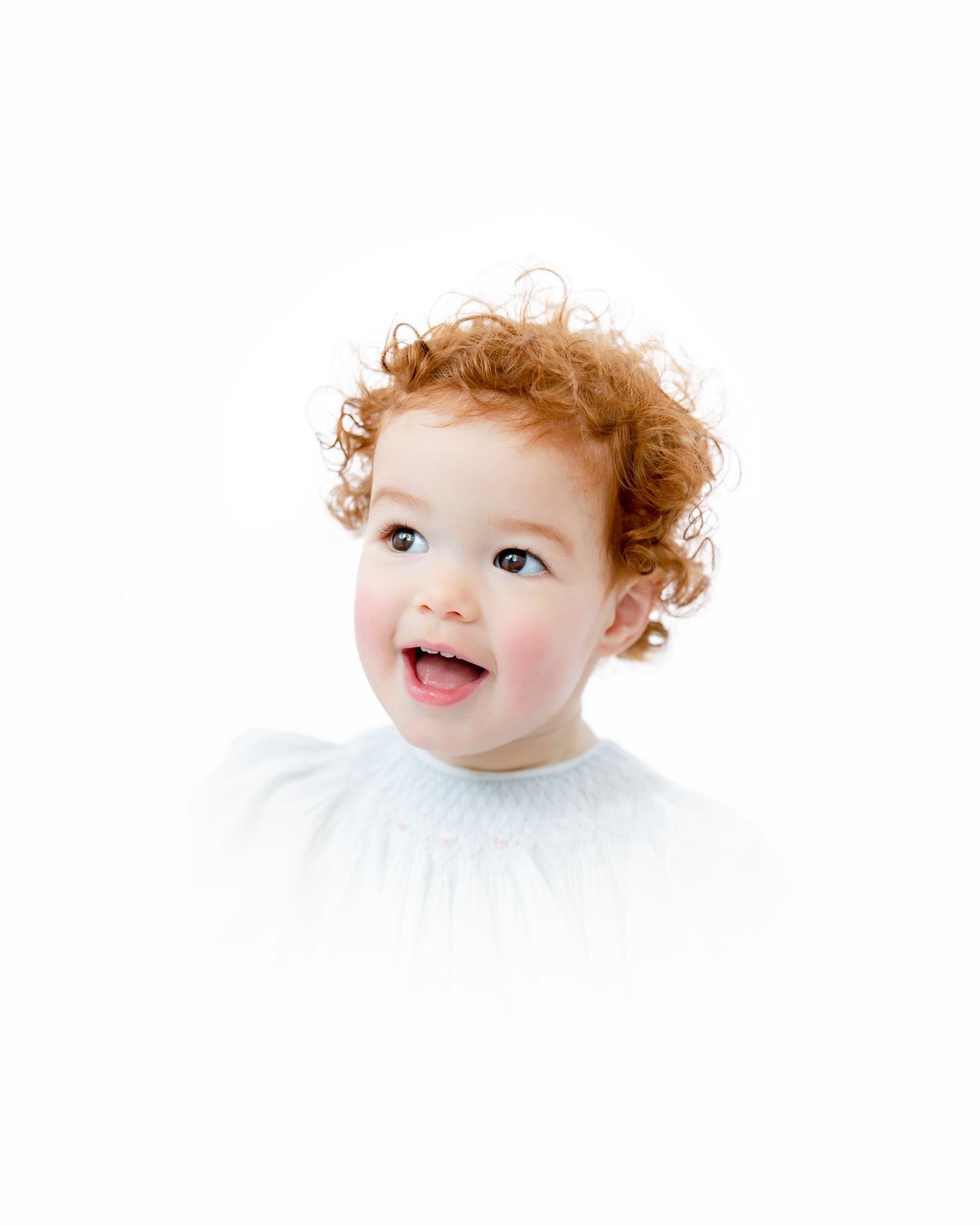 A laughing toddler with ginger curls dressed in white on a bright white backdrop.