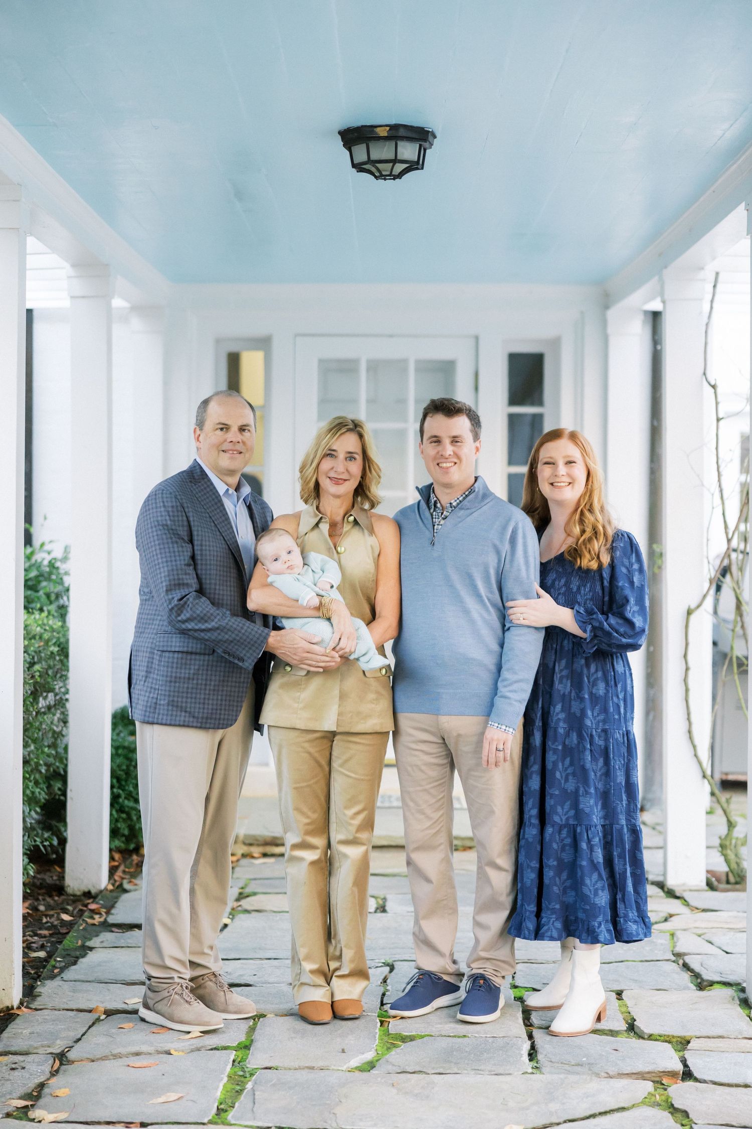 A family poses together on a white porch while holding a newborn baby.