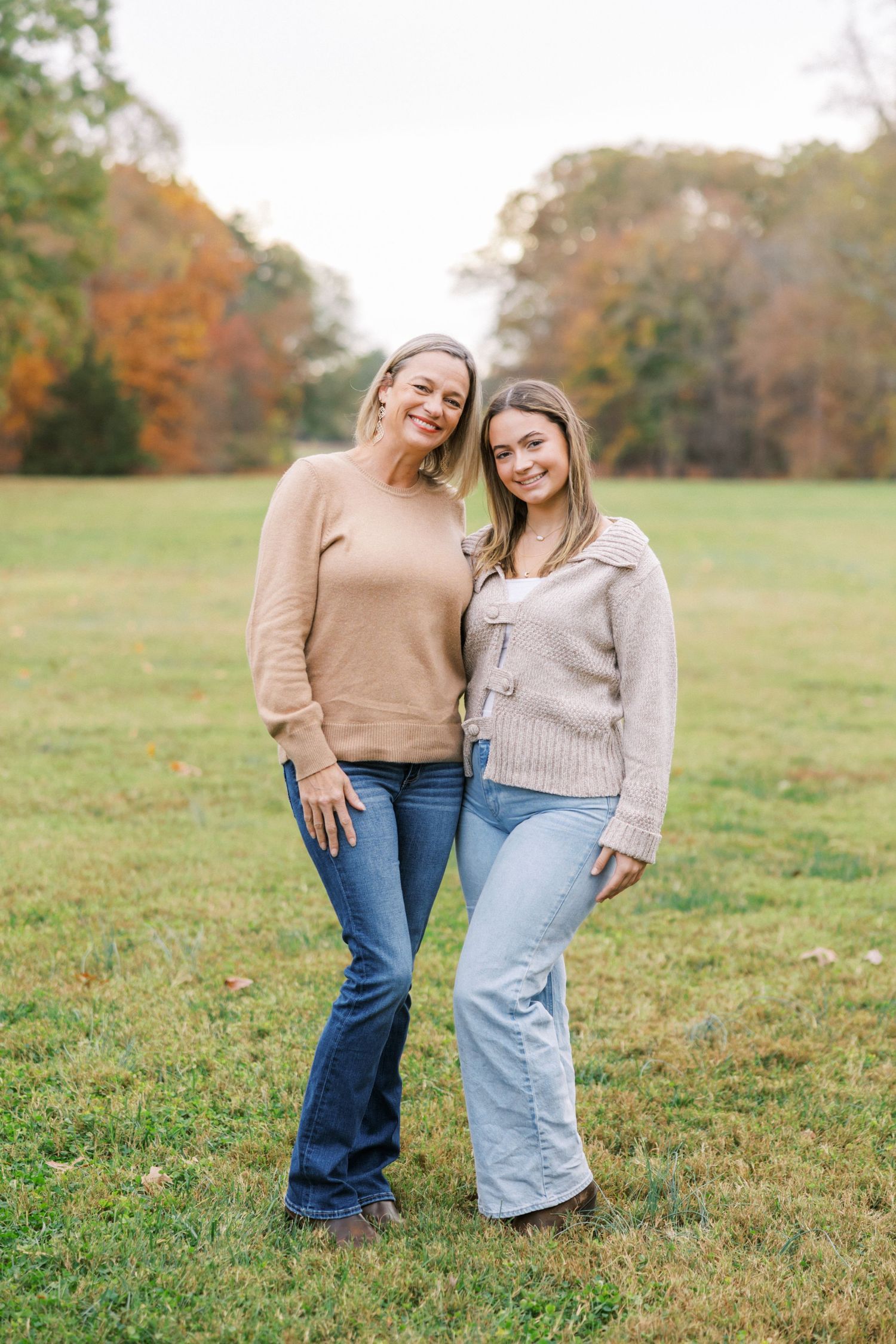Two friends wearing cozy sweaters pose together on an autumn field with fall foliage in the background.