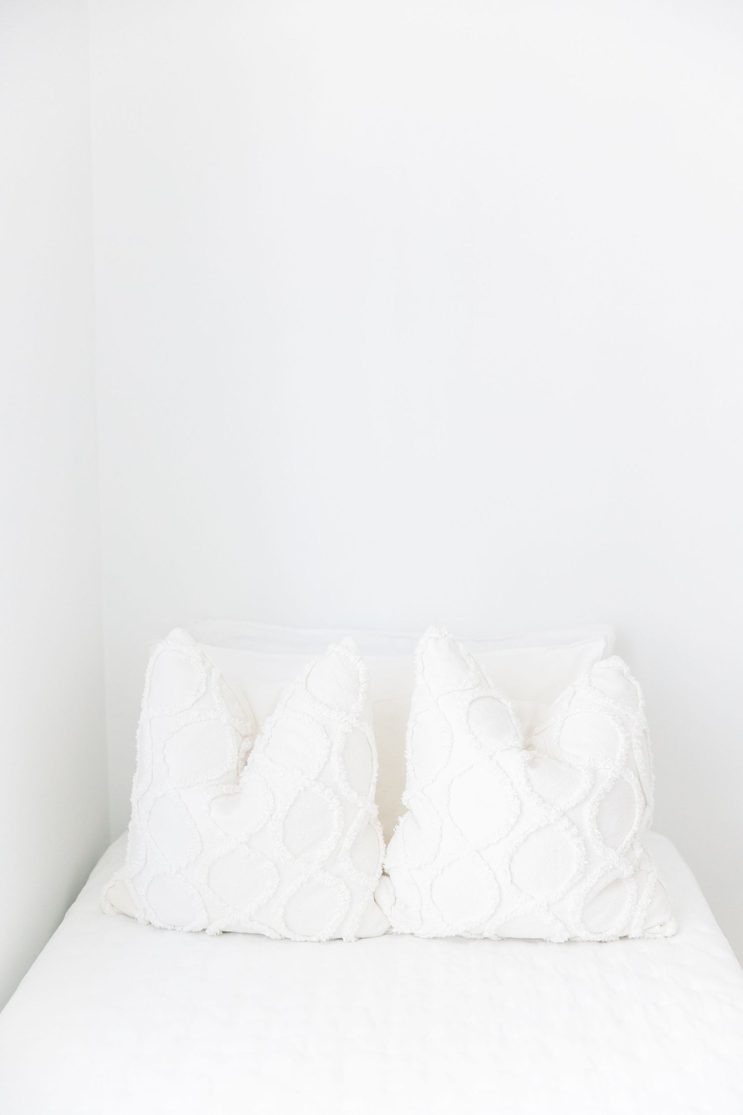 Two white fluffy pillows on a bed with white bedding against a plain white wall background.