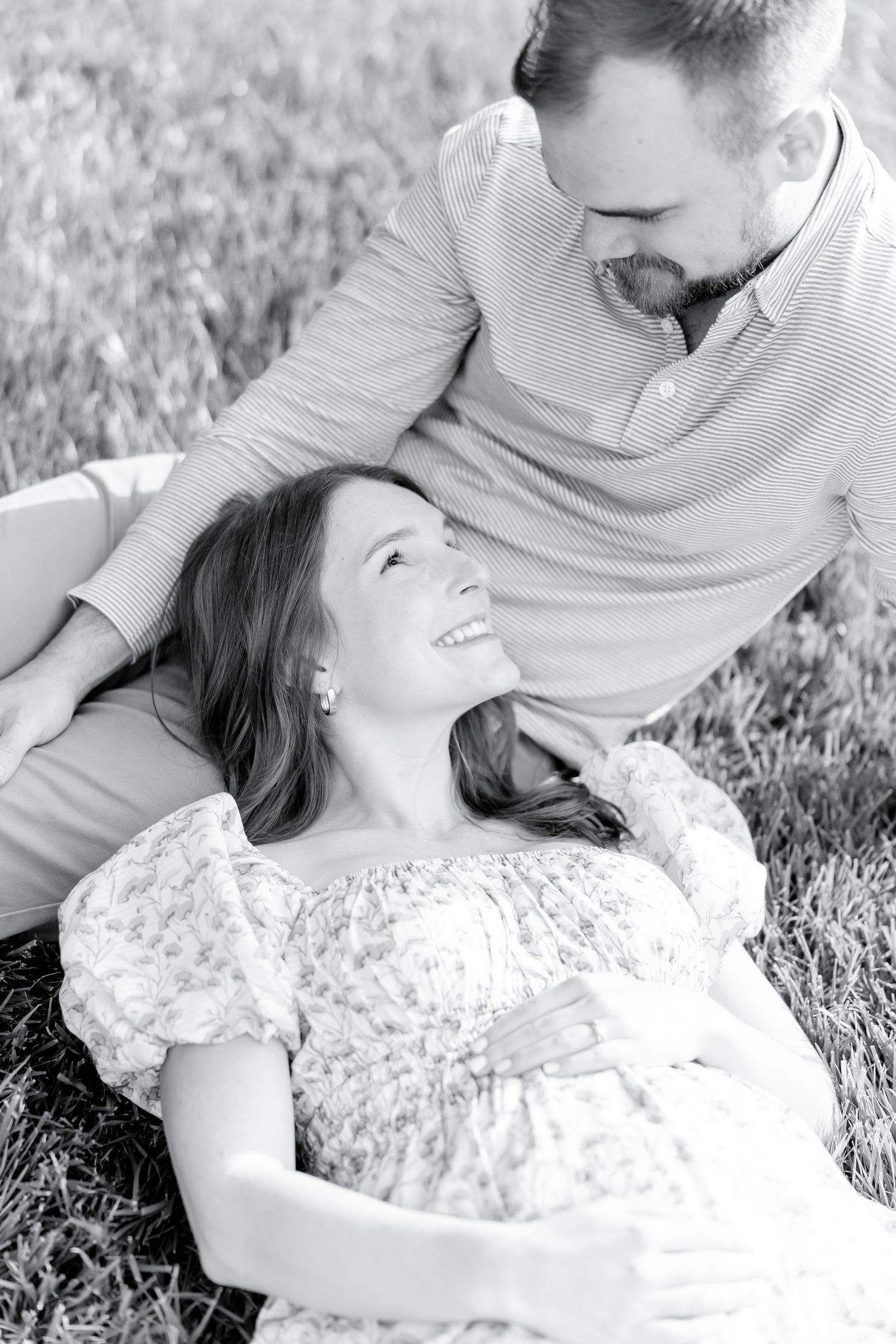 Black and white portrait of a romantic couple resting in grass, with woman laying on her back in lace dress smiling.