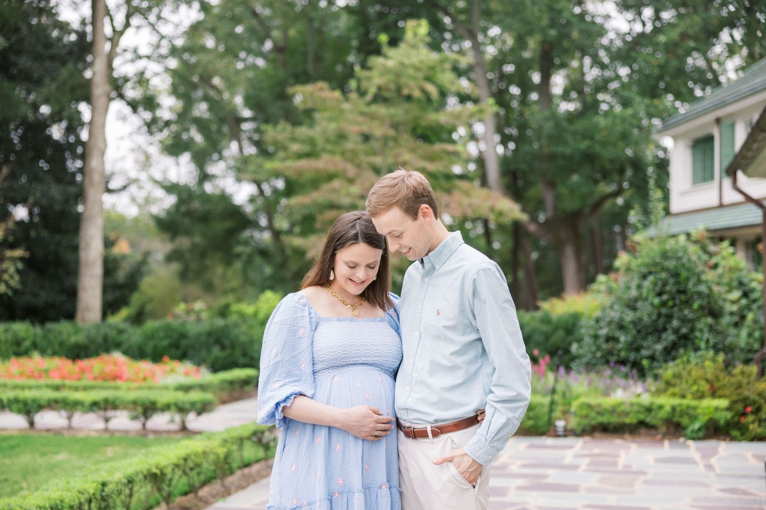 Expecting couple stands together in a garden setting wearing light blue and white clothing during a maternity photoshoot.