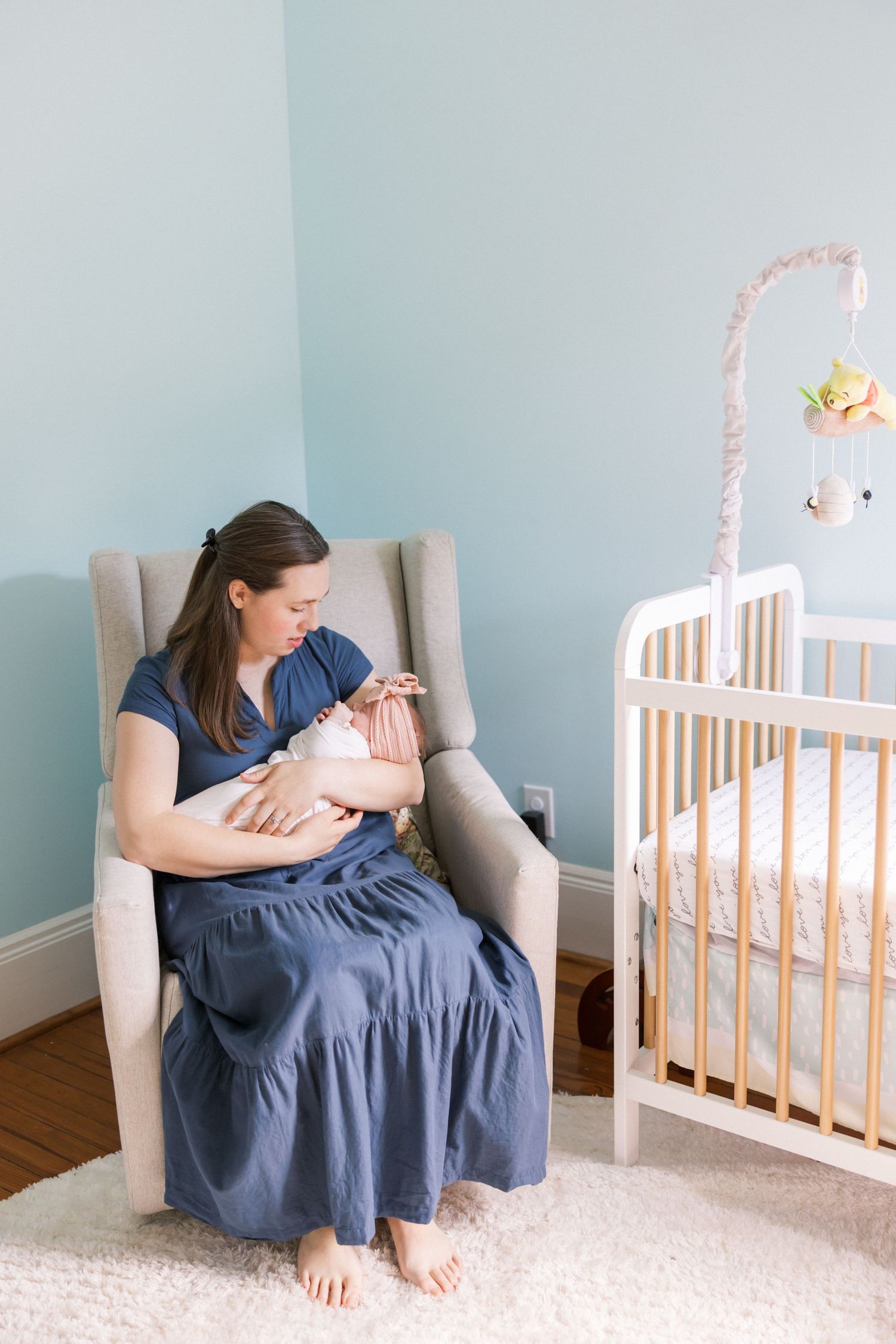 Mother in navy dress sitting in gray armchair holding sleeping newborn next to white crib.