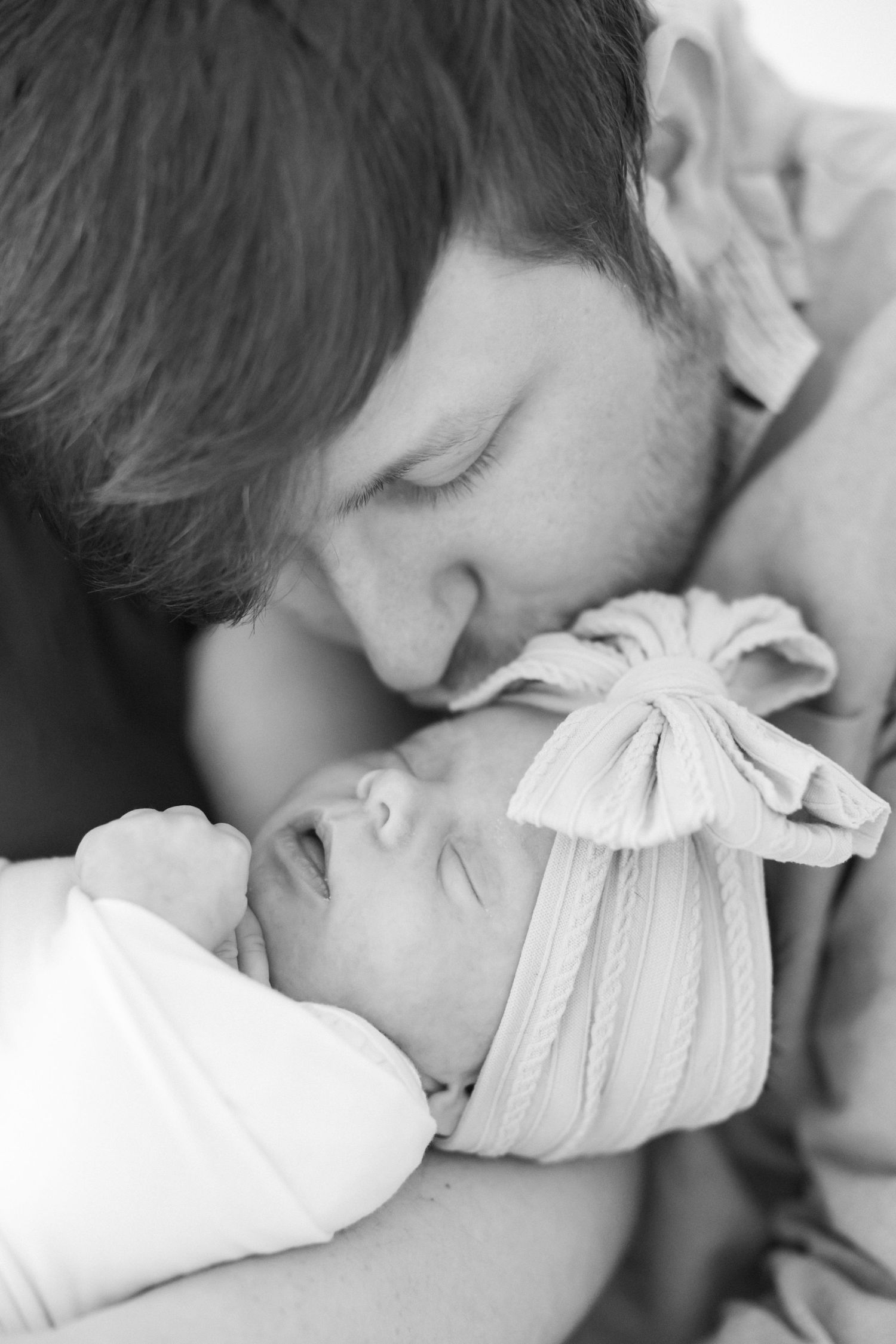 Close-up black and white portrait of parent tenderly kissing newborn with bow headband.