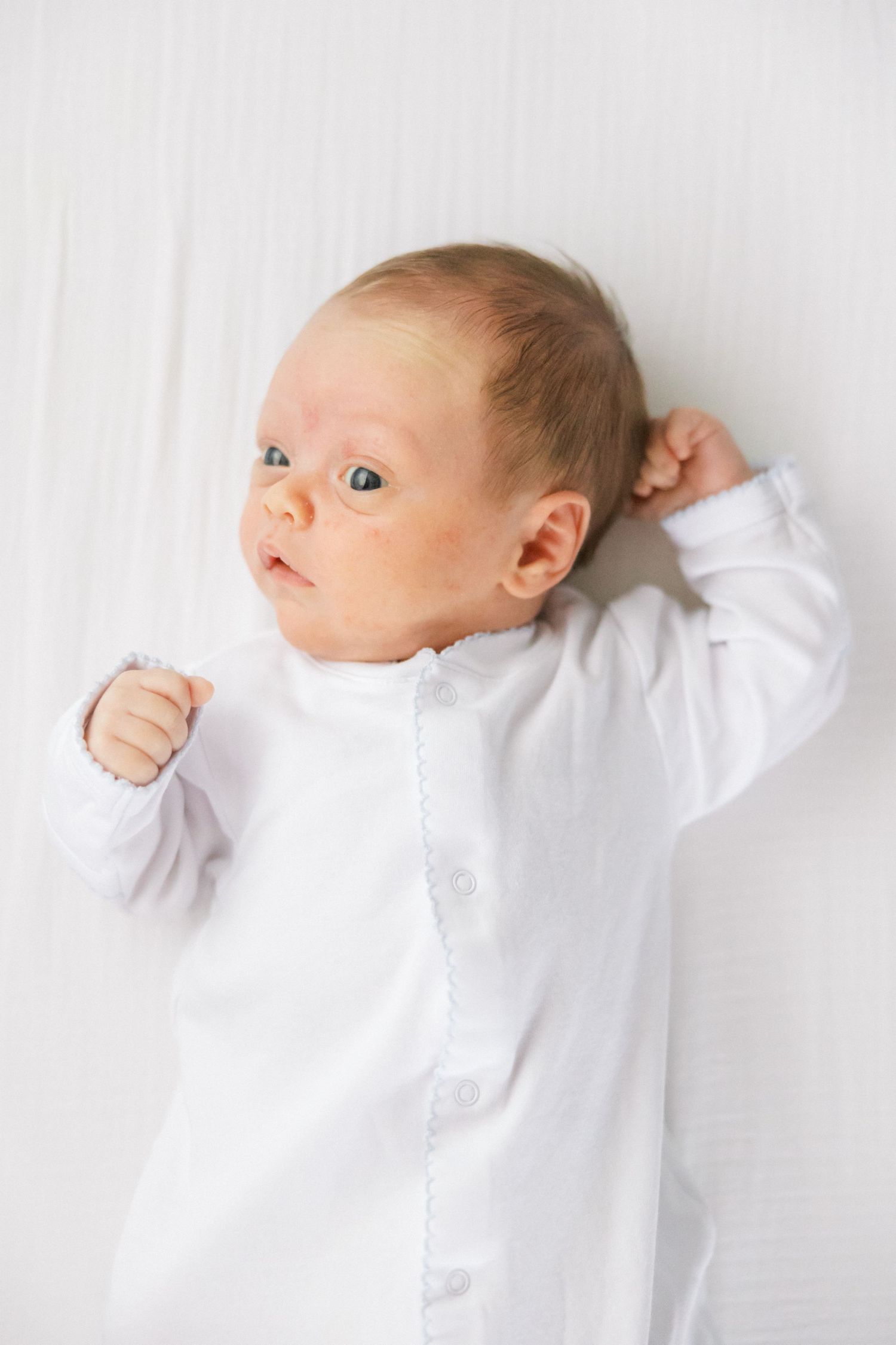 Alert newborn baby in white onesie lying on white background looking at camera.