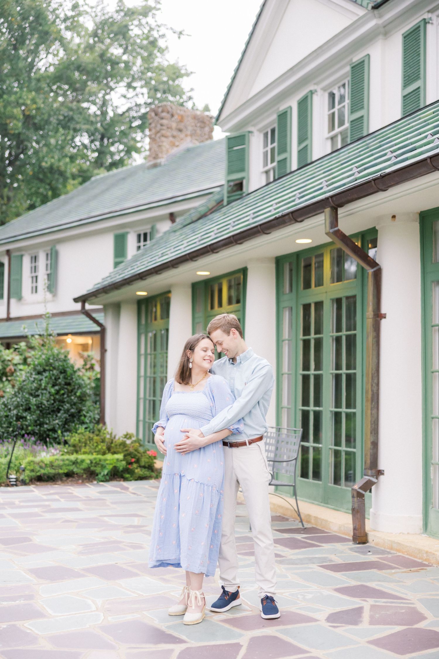 Couple embraces in front of classic white house with green shutters and stone pathway.