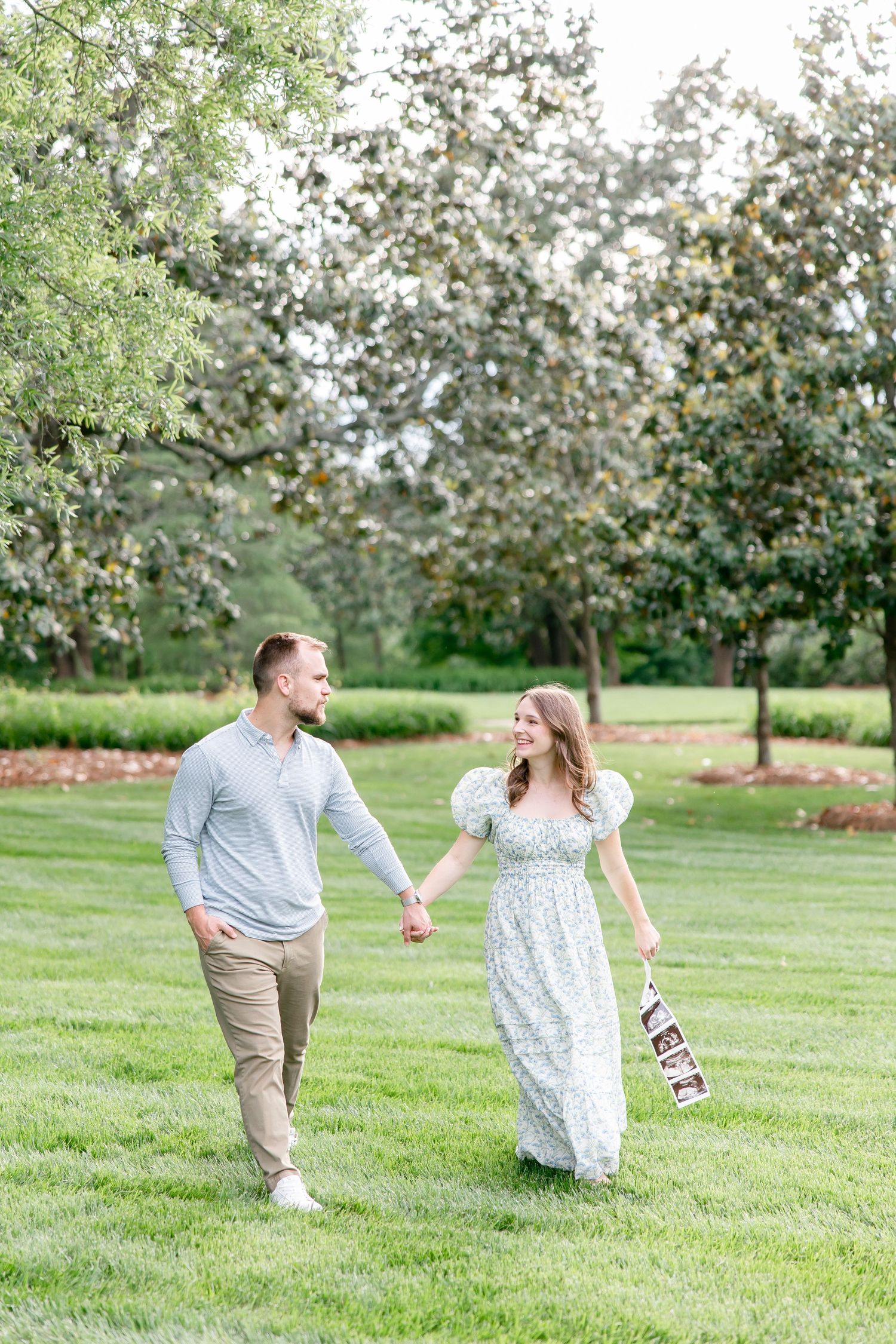 A romantic couple walks hand in hand through a lush garden with blooming trees on a bright summer day.