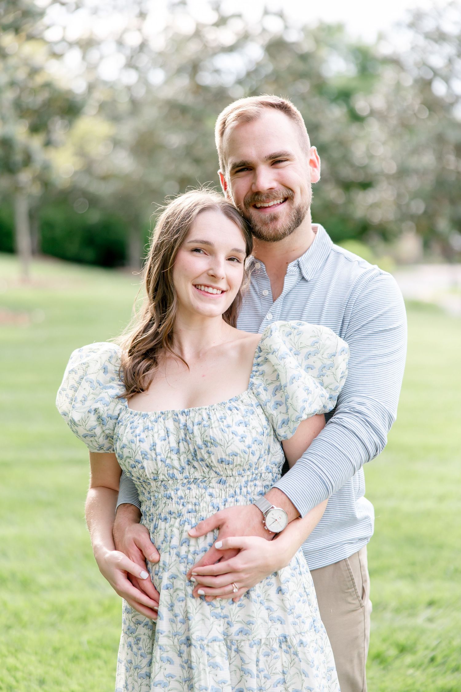 A couple embraces outdoors in a grassy field, the woman wearing a floral dress while sharing a tender moment together.