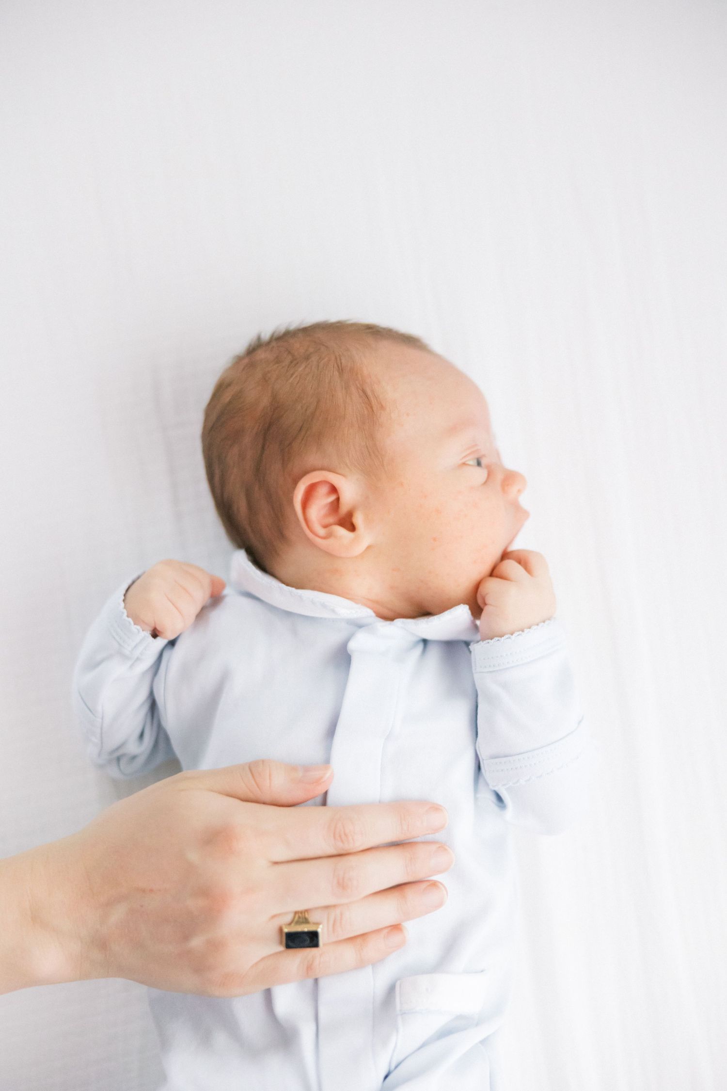 A newborn baby in light blue clothing looks to the side while being held against white background.