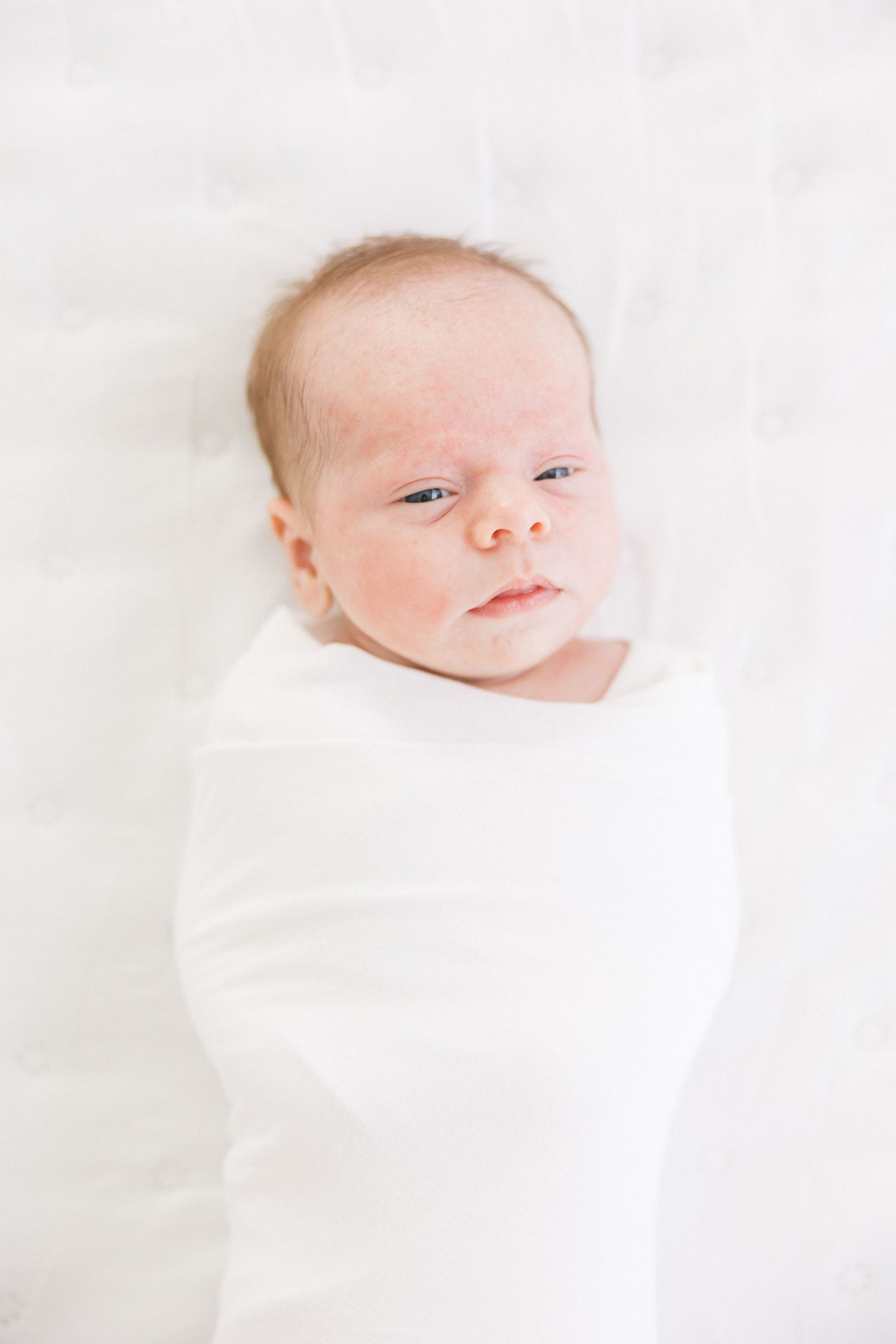 Alert newborn swaddled in white fabric gazes at camera against soft white background.