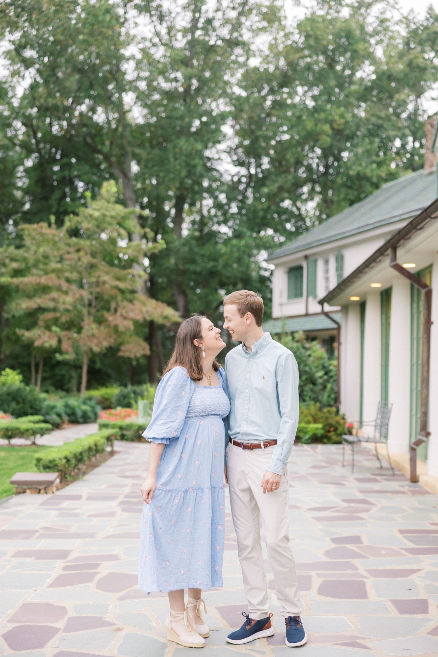 A couple in coordinating blue and white outfits share a tender moment on a stone patio outside a classic home.