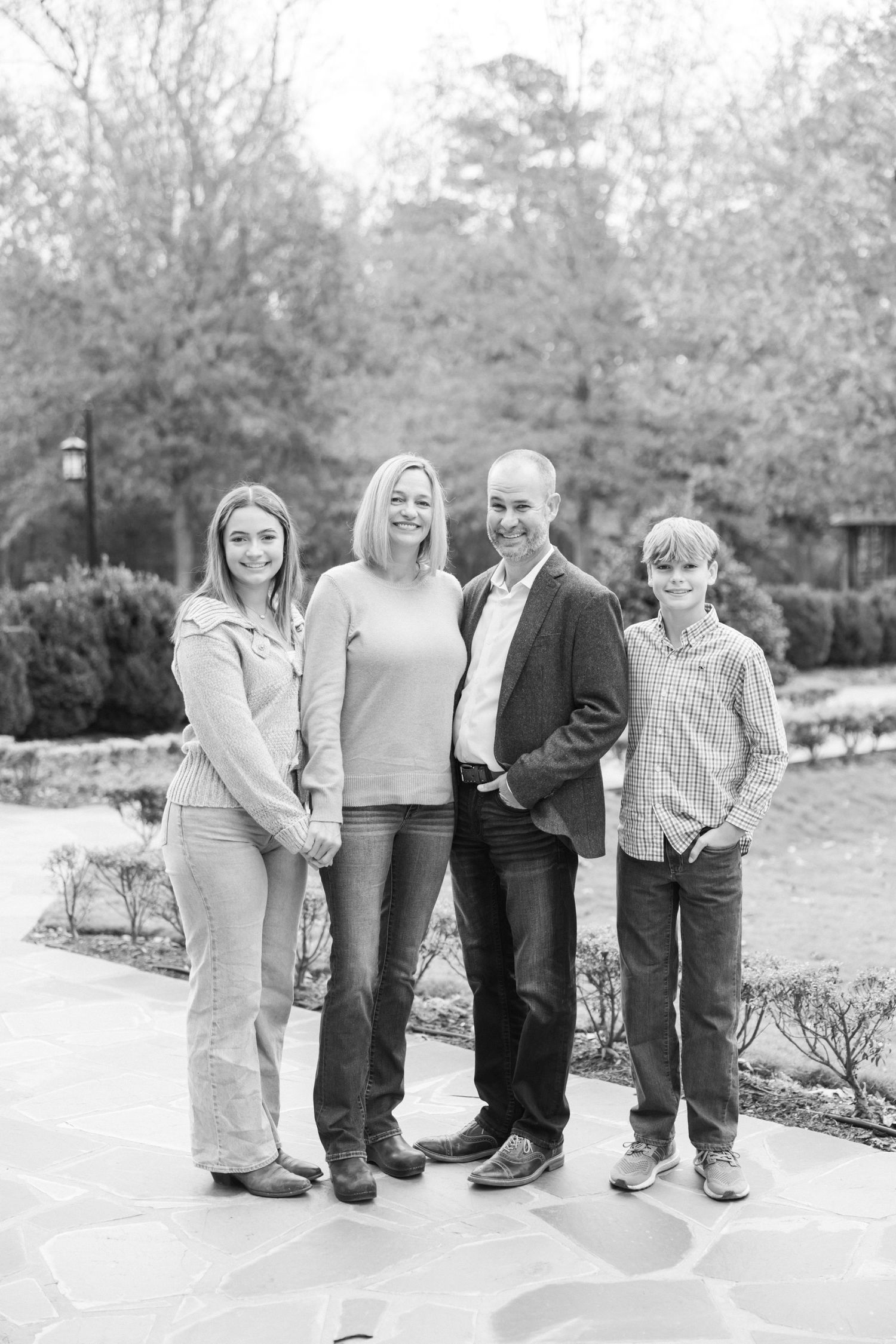 A black and white portrait of a family of four standing together outdoors on a stone patio surrounded by landscaping.